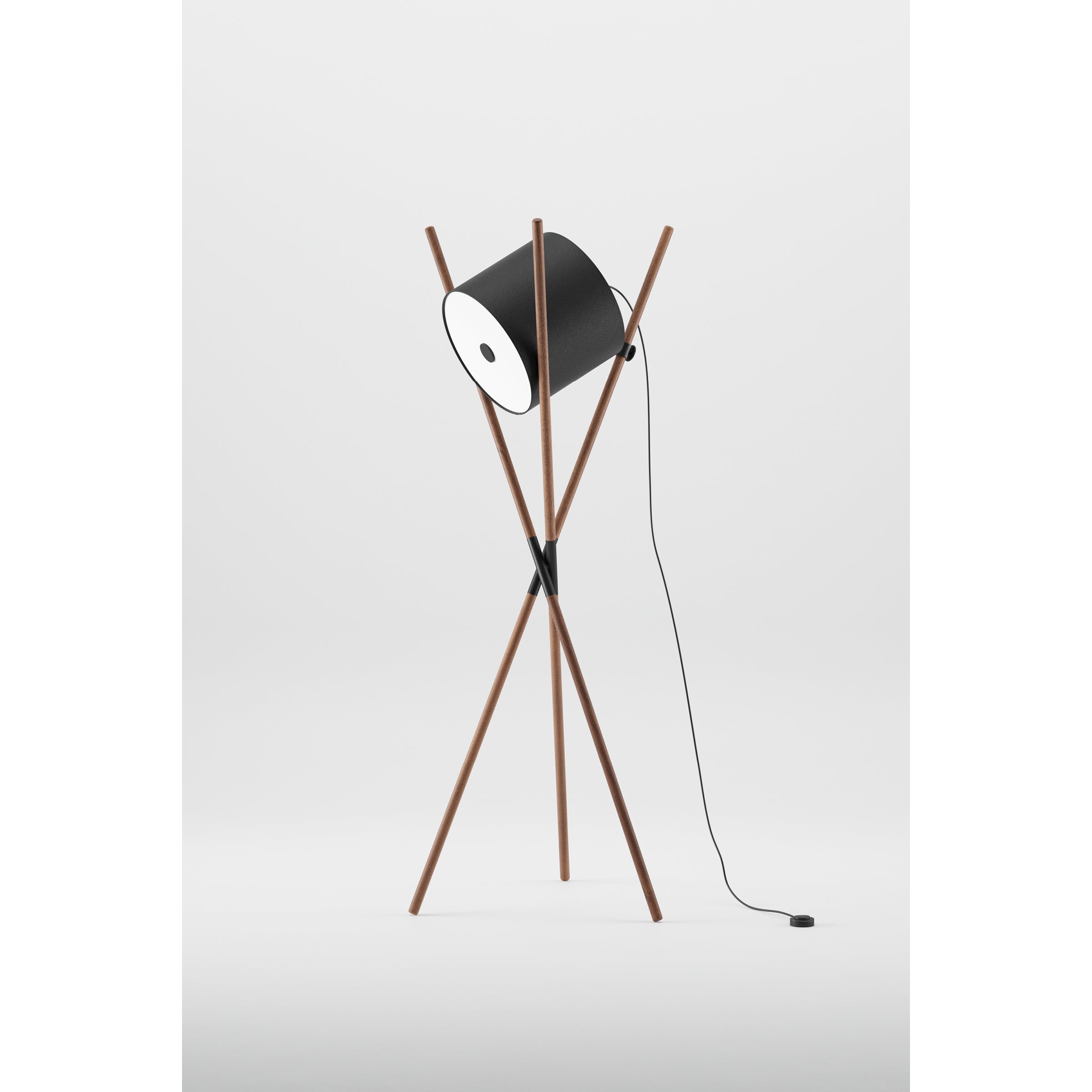 The Artisan SHIFT Lamp boasts a minimalist elegance with its black cylindrical shade on wooden tripod struts. Its flat-pack design and sleek black power cord complement its modern style against a white backdrop.