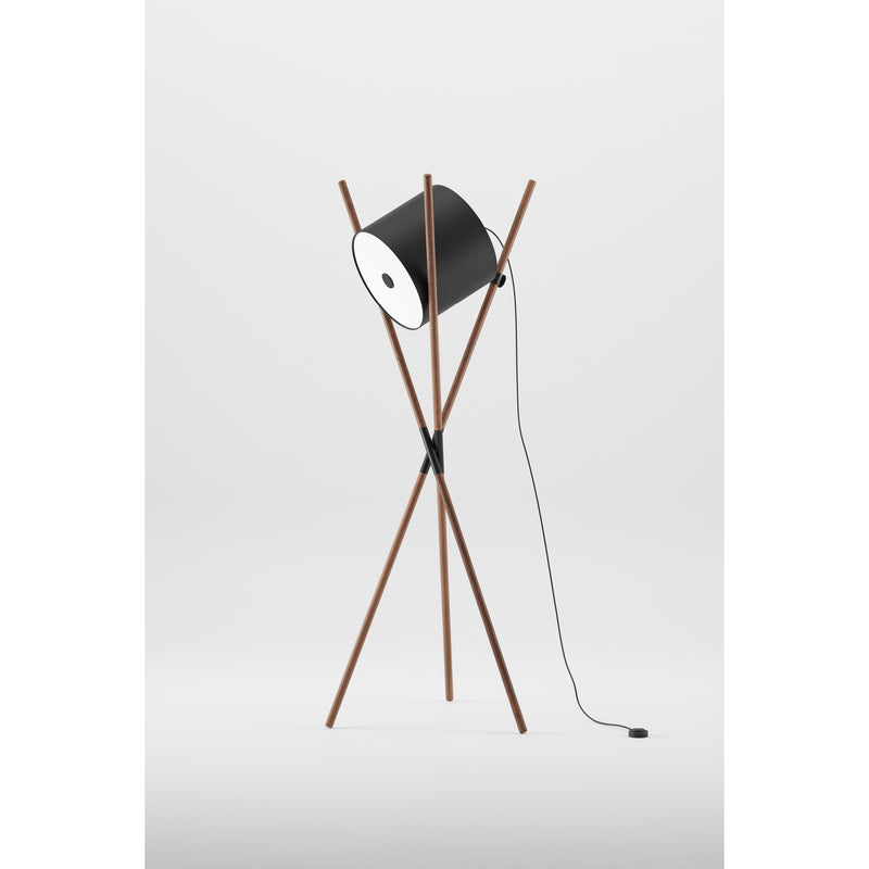 The Artisan SHIFT Lamp boasts a minimalist elegance with its black cylindrical shade on wooden tripod struts. Its flat-pack design and sleek black power cord complement its modern style against a white backdrop.