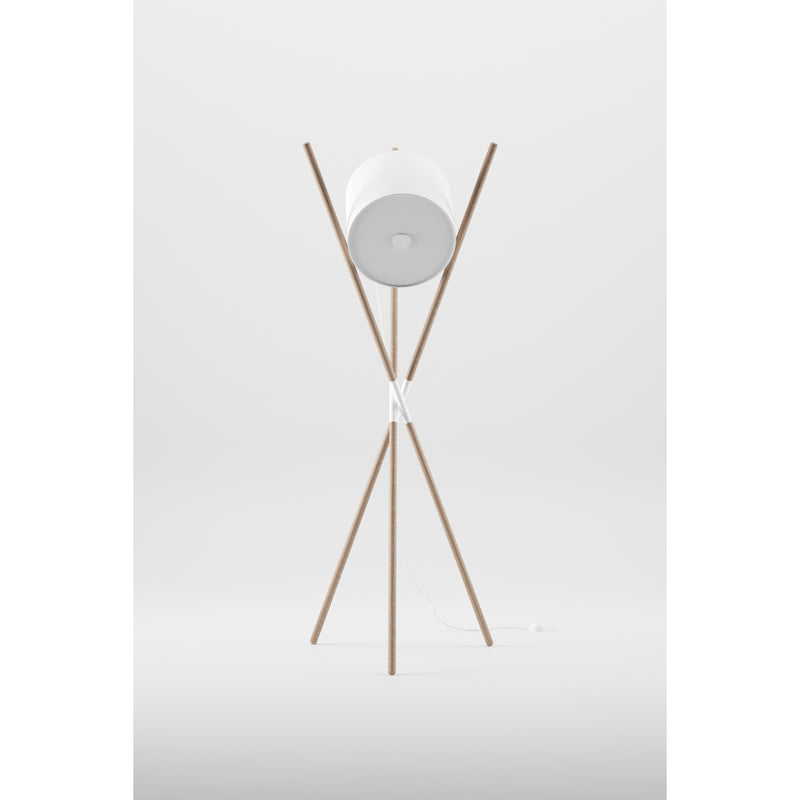 The SHIFT Lamp by Artisan is a modern floor lamp with a flat-pack design featuring a tripod base made of wooden struts, a cylindrical white lampshade, and intersecting poles for balance. A white cord elegantly rests at the base, complementing its minimalist style.