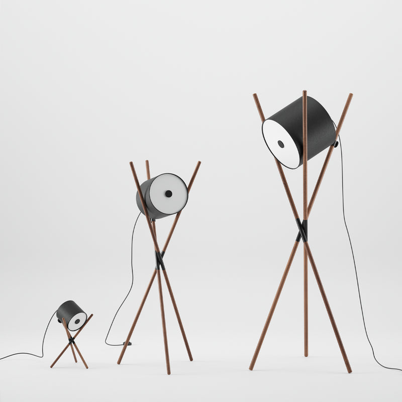 Three Artisan SHIFT wooden tripod lamps of varying heights, featuring a flat-pack design with cylindrical black shades, stand in a row on a white background. Each lamp has a cord hanging down, and the shades are positioned at different angles.