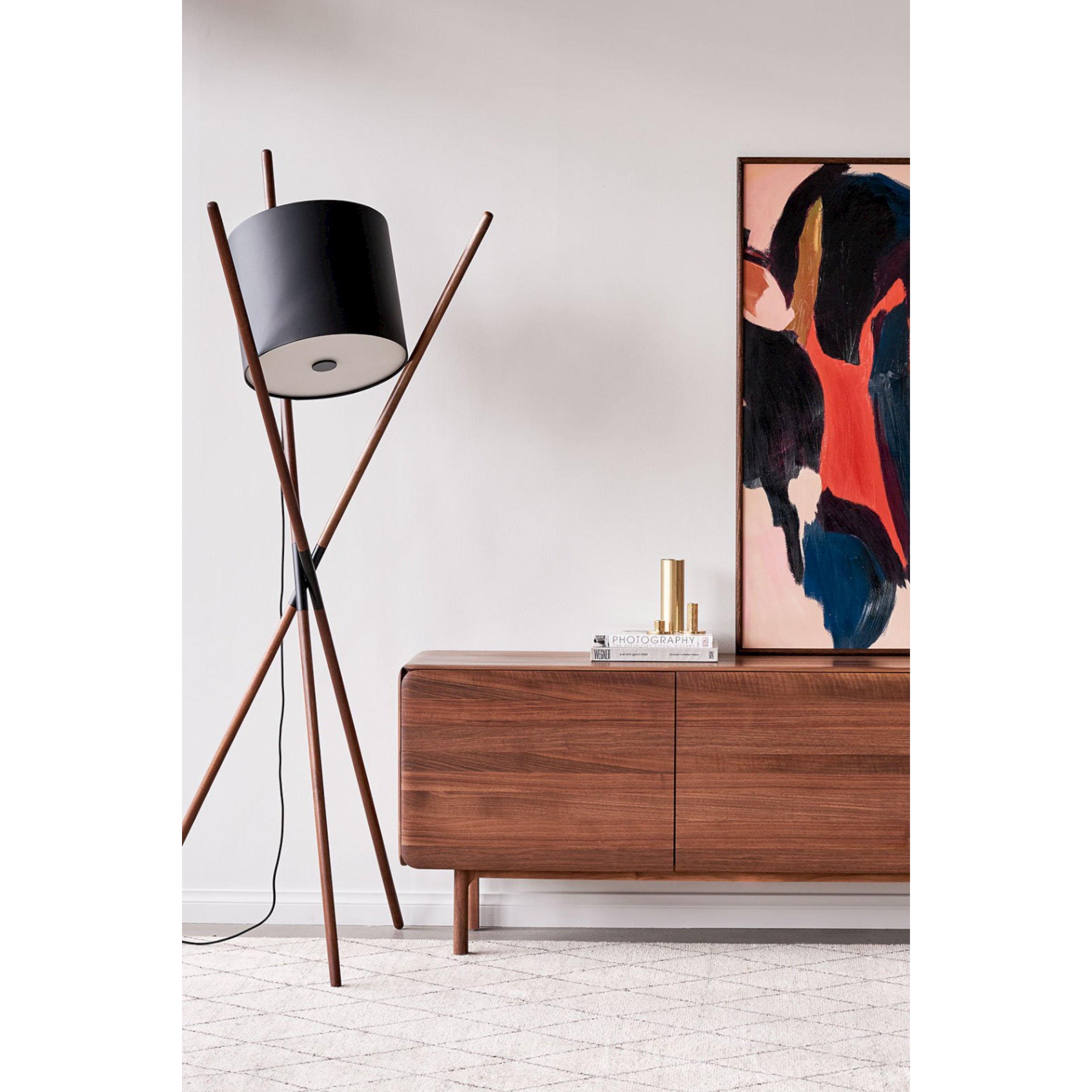 A modern living room scene showcases a sleek wooden sideboard adorned with books and a brass vase, reflecting flat-pack design. To the left stands Artisans SHIFT Lamp featuring a black shade, while an abstract art piece bursting with bold colors is displayed above the sideboard.