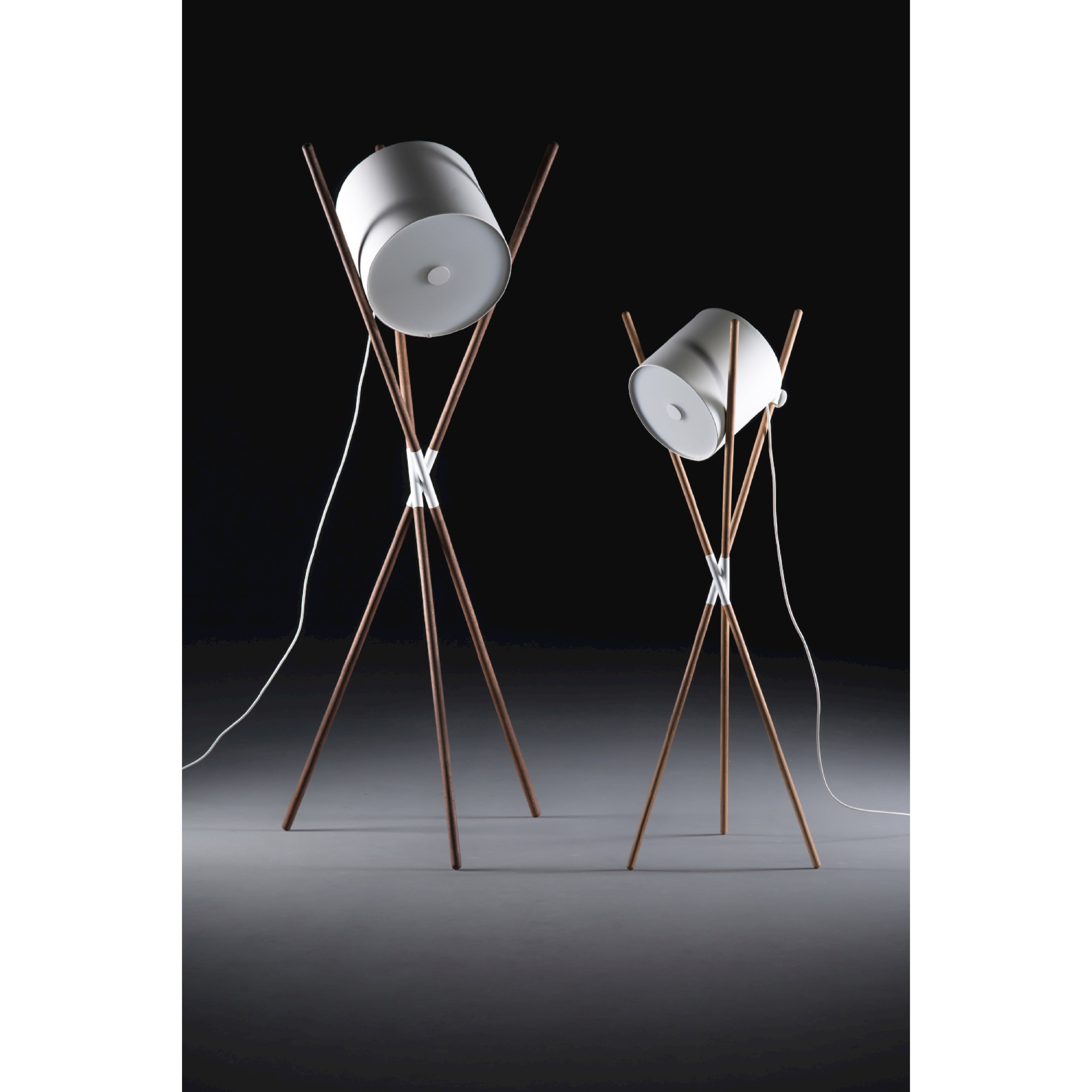 Two Artisan SHIFT Lamps feature white cylindrical shades on wooden tripod stands with sleek struts at varying heights. The lamps have a flat-pack design and are set against a dark background with visible white power cords.