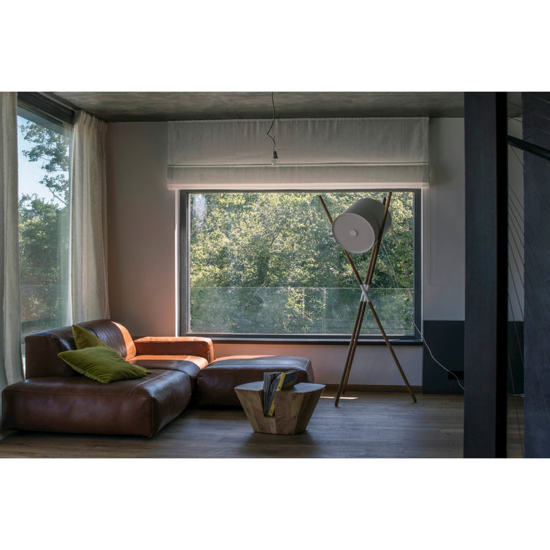 A cozy living room showcases a brown leather sectional adorned with green pillows. A flat-pack coffee table with books adds charm, while Artisans SHIFT Lamp, featuring a large white shade, delivers a warm glow. Natural light enhances the space through the window with lush garden views.