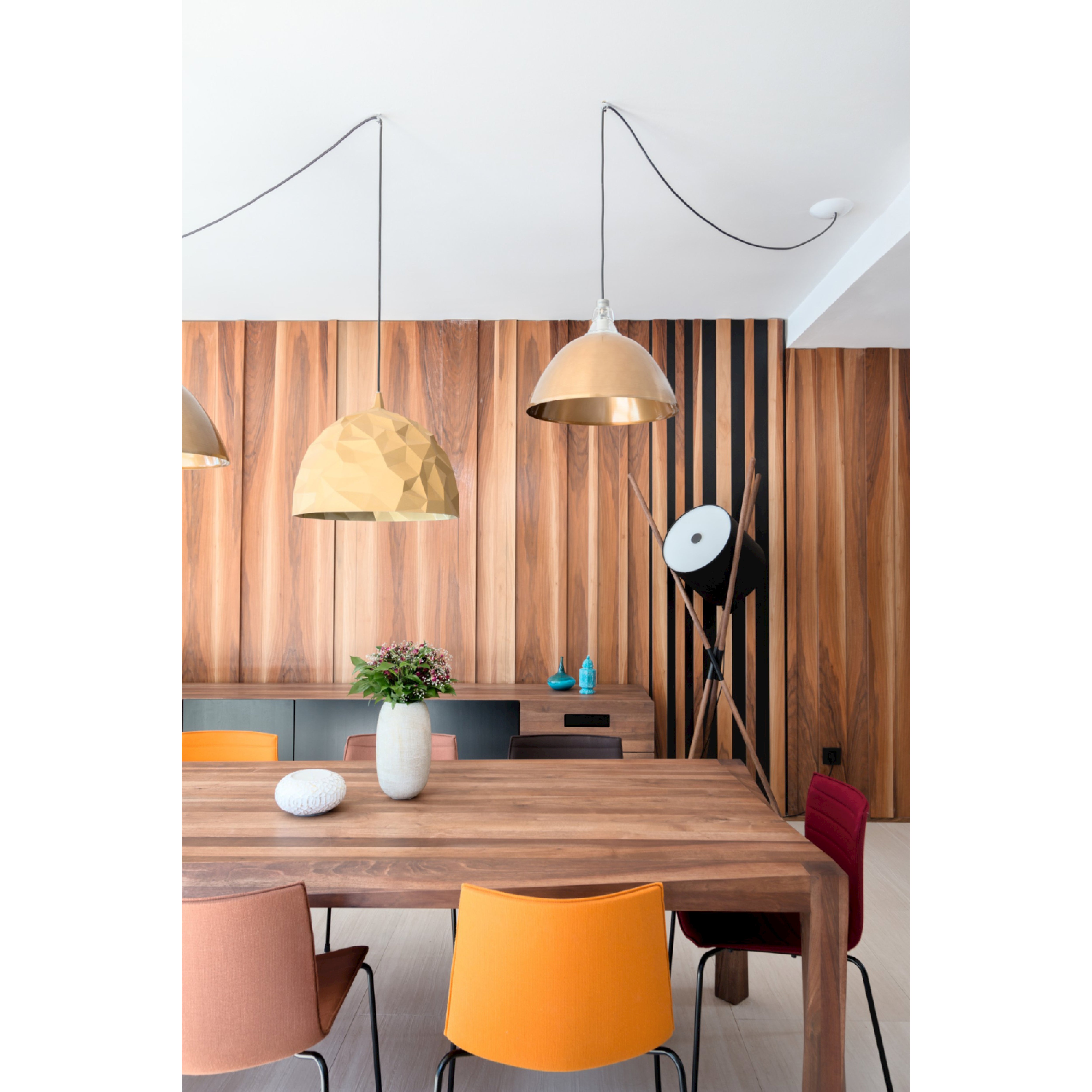 A modern dining space with a flat-pack wooden table, multicolored chairs, and two geometric gold pendant lights. The textured wall has black vertical stripes like wooden struts, while a sideboard with vases and greenery is brightened by the Artisan SHIFT Lamp.