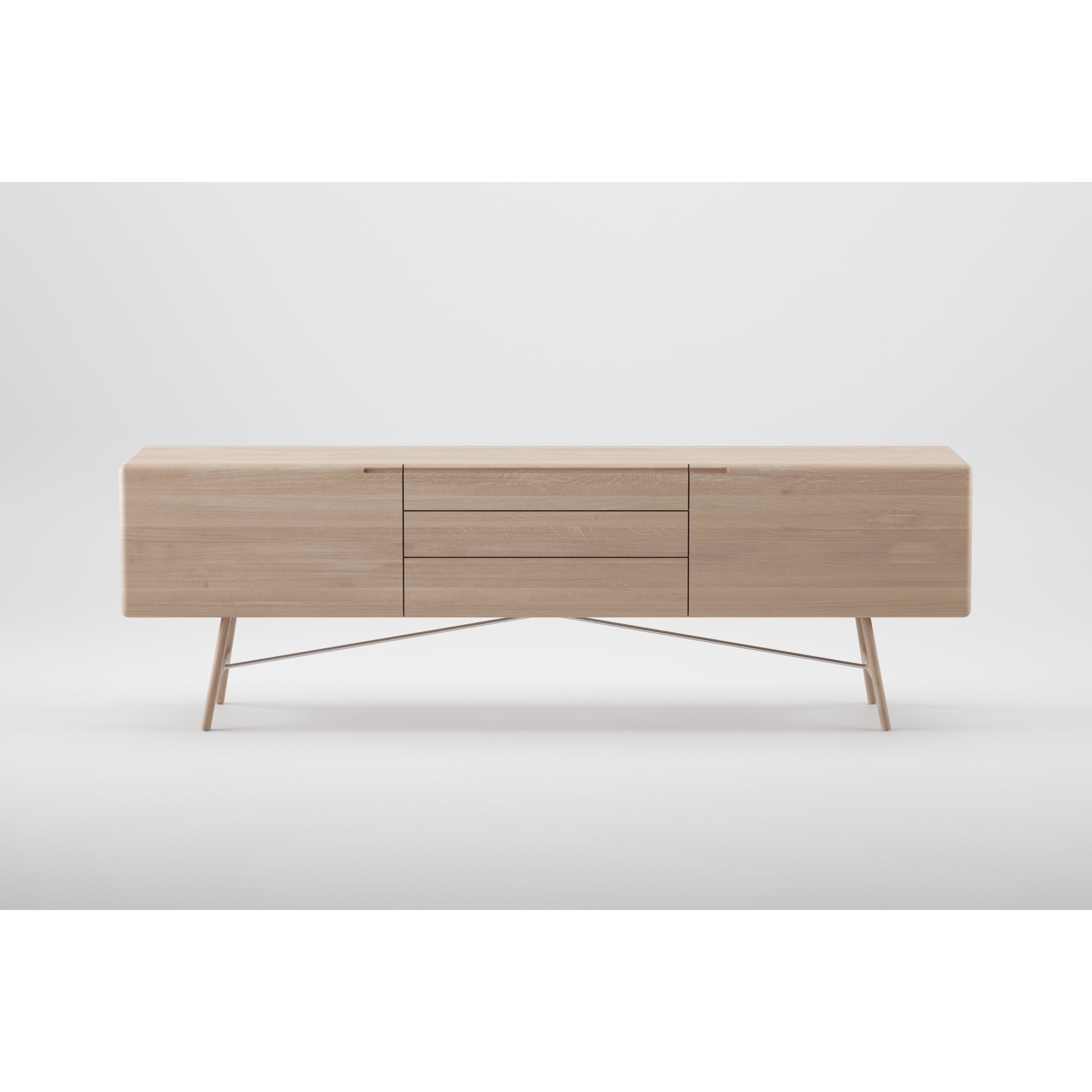 The TESA Sideboard by Artisan is a minimalist piece crafted from solid wood with a light finish. It features two side cabinets, three central drawers, and angled legs connected by a crossbar. It stands against a plain white background and is finished with natural oils.
