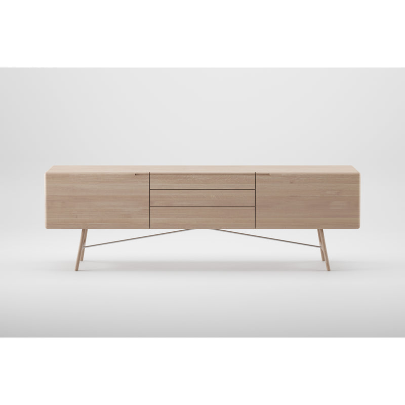 The TESA Sideboard by Artisan is a minimalist piece crafted from solid wood with a light finish. It features two side cabinets, three central drawers, and angled legs connected by a crossbar. It stands against a plain white background and is finished with natural oils.