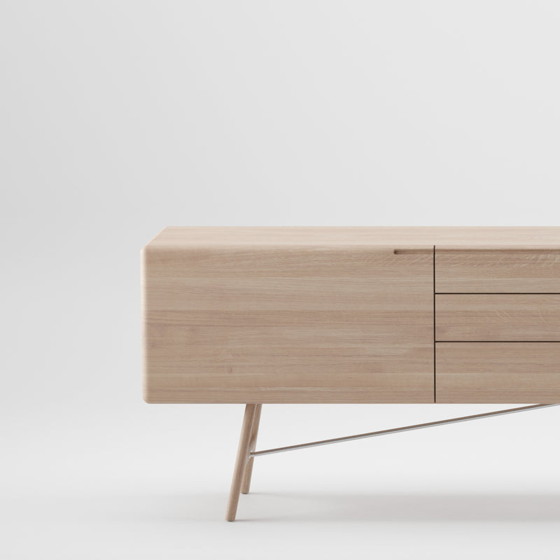 The Artisan TESA Sideboard is a minimalist solid wood piece set against a white background. It showcases two doors, three drawers on sleek angled legs with a metal support rod, and is finished with natural oils for eco-friendliness.