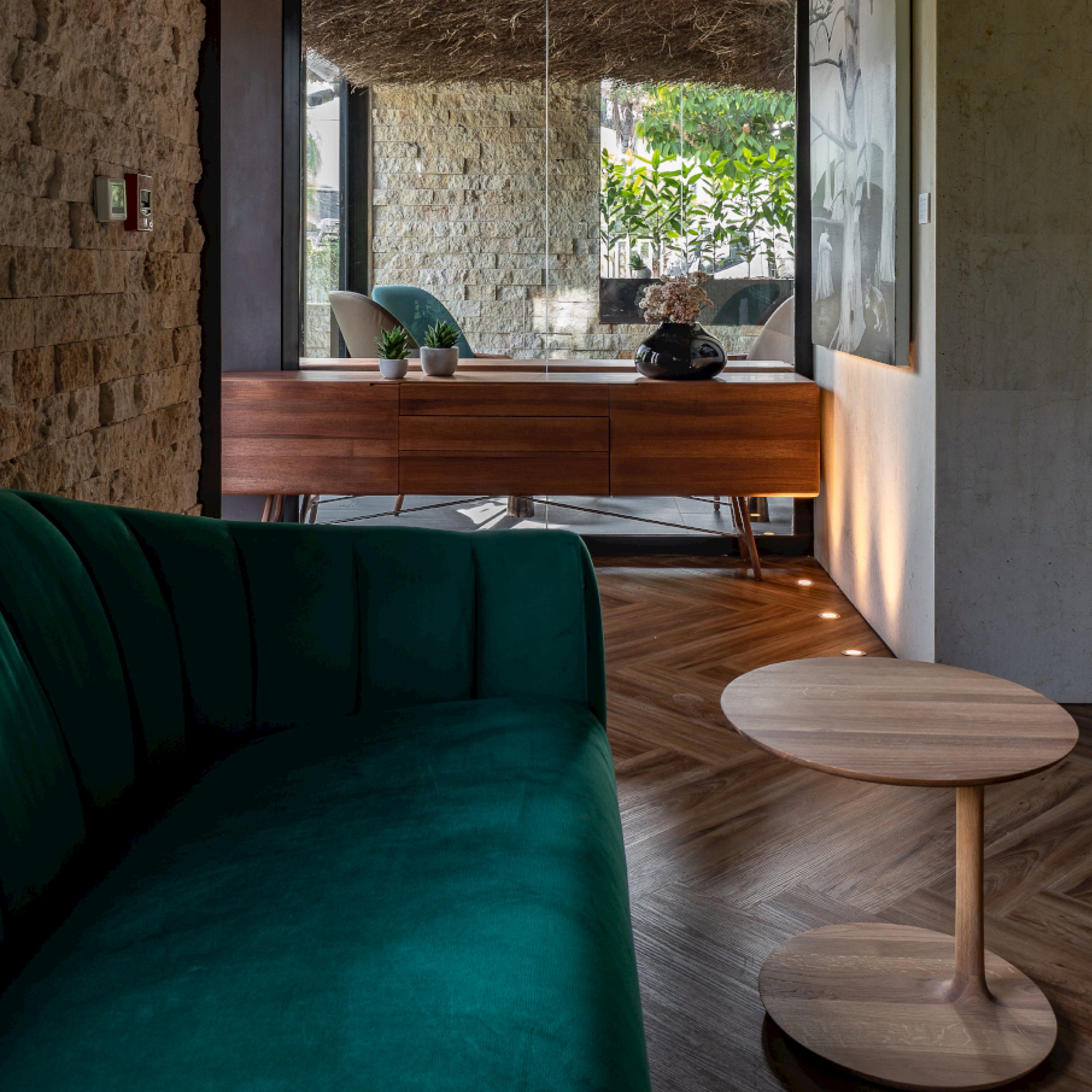 A cozy living space showcases a dark green sofa and a round solid wood coffee table on a herringbone-patterned floor. In the background, the environmentally-friendly TESA Sideboard by Artisan holds a teapot and decor next to glass doors opening to a sunny outdoor area.