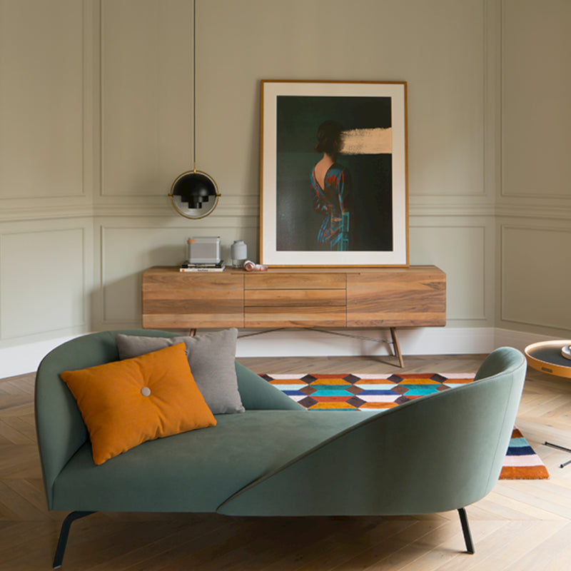 Modern living room with a green chaise lounge and orange-gray pillows. A colorful geometric rug covers the floor. The Artisan TESA Sideboard, made from eco-friendly materials, displays a framed abstract painting, while a black pendant light completes the decor.