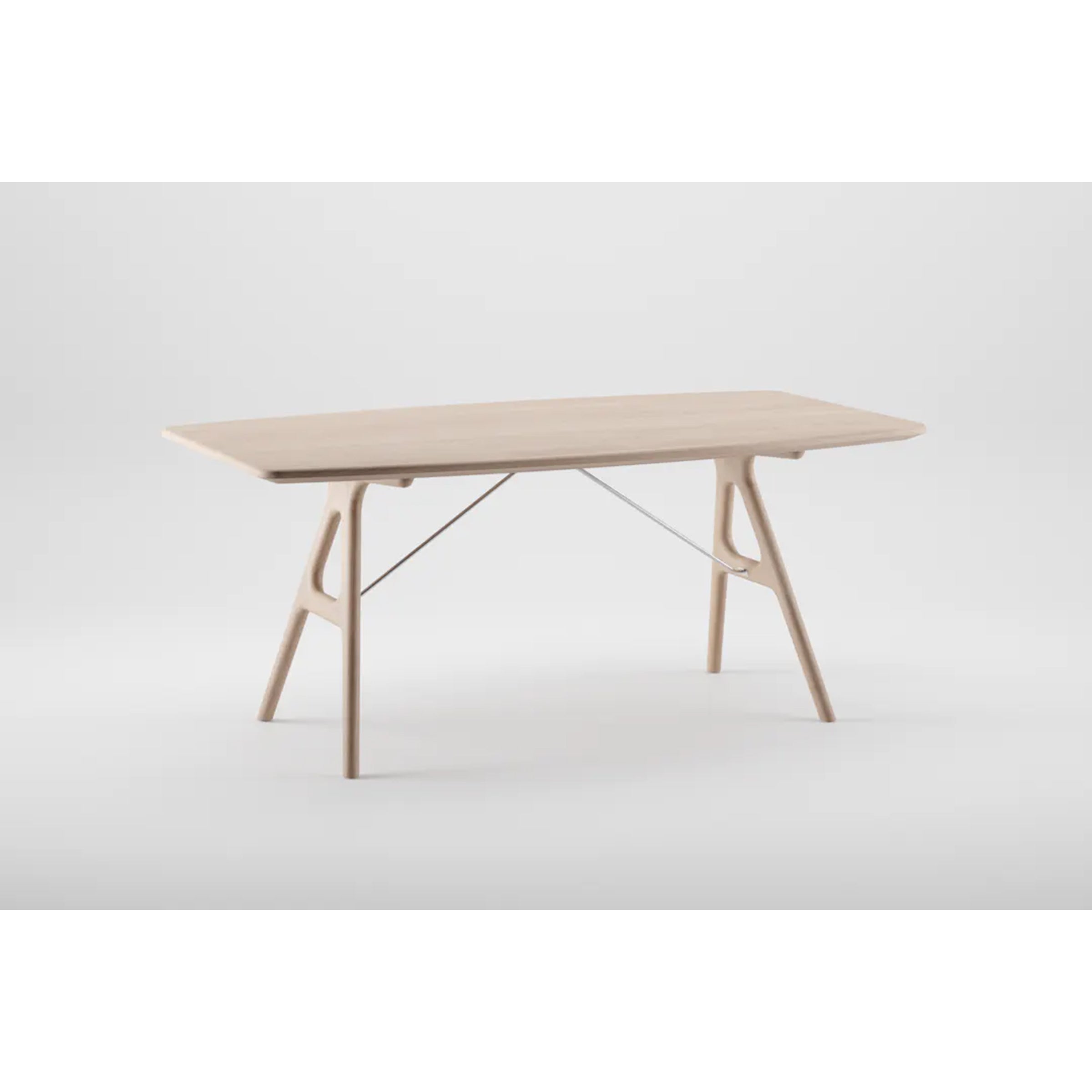 The TESA Table by Artisan is a modern rectangular piece crafted from eco-friendly solid wood, featuring a light finish, sleek angled legs, and a subtle metal support bar. It stands elegantly on a plain white background.