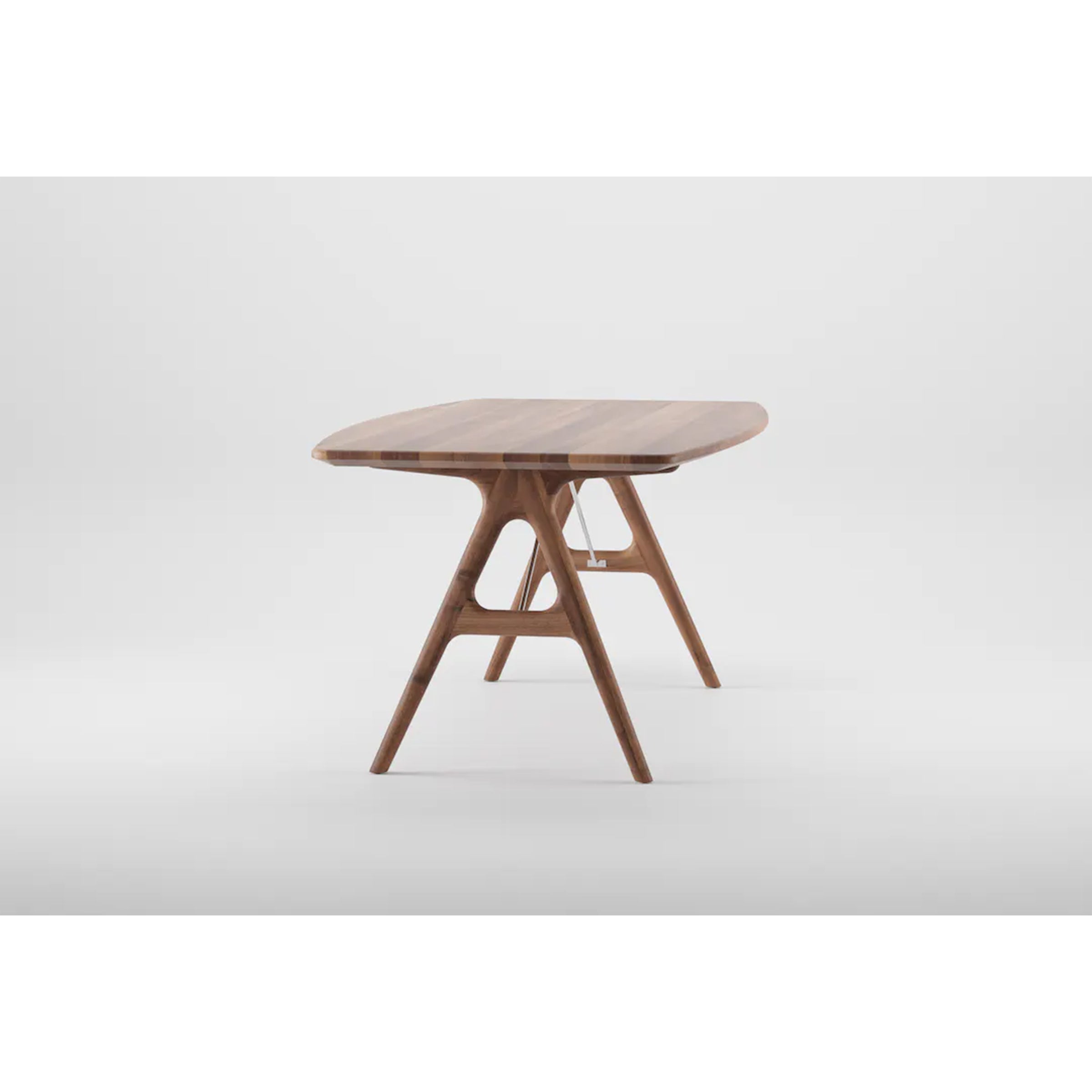 The TESA Table by Artisan features a modern angular design crafted from solid wood. It boasts a rounded-edged tabletop supported by crossed wooden legs linked with a horizontal beam, finished with natural oils, and set against a plain white background to emphasize its sustainable charm.