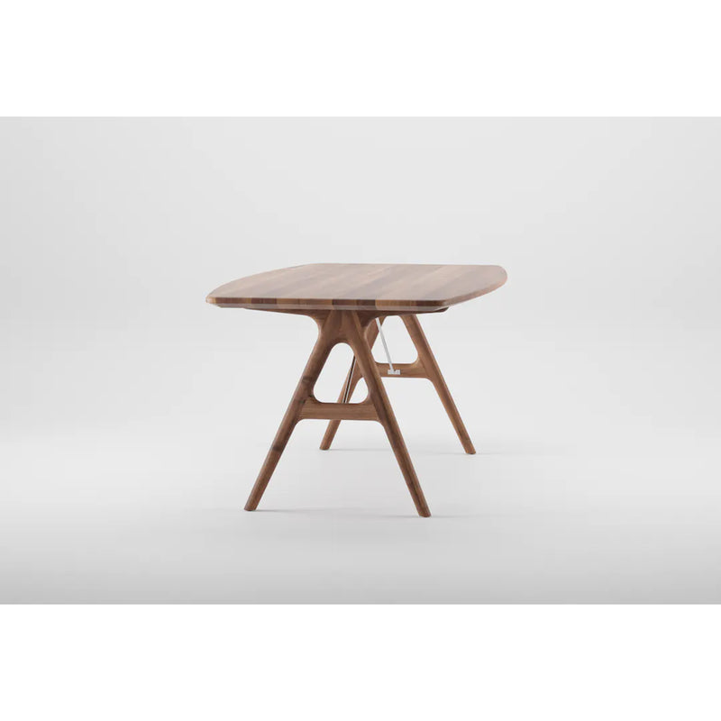 The TESA Table by Artisan features a modern angular design crafted from solid wood. It boasts a rounded-edged tabletop supported by crossed wooden legs linked with a horizontal beam, finished with natural oils, and set against a plain white background to emphasize its sustainable charm.