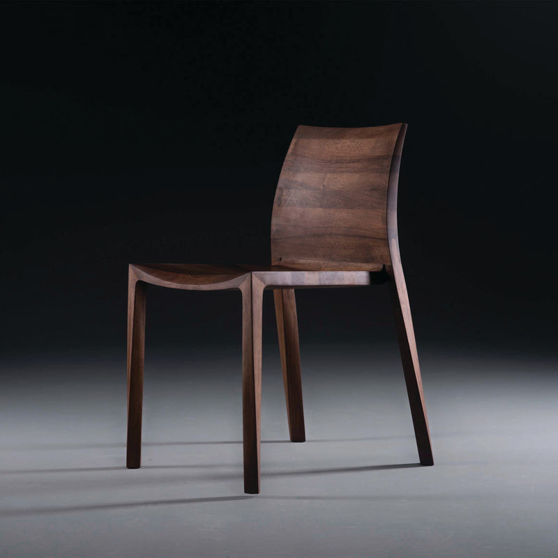 The TORSIO Chair by Artisan features a sleek, modern design with a solid wood construction. Its curved backrest and slightly angled legs are shown against a dark background. The chairs smooth finish, made with natural oils, highlights its minimalist design and eco-friendly commitment.