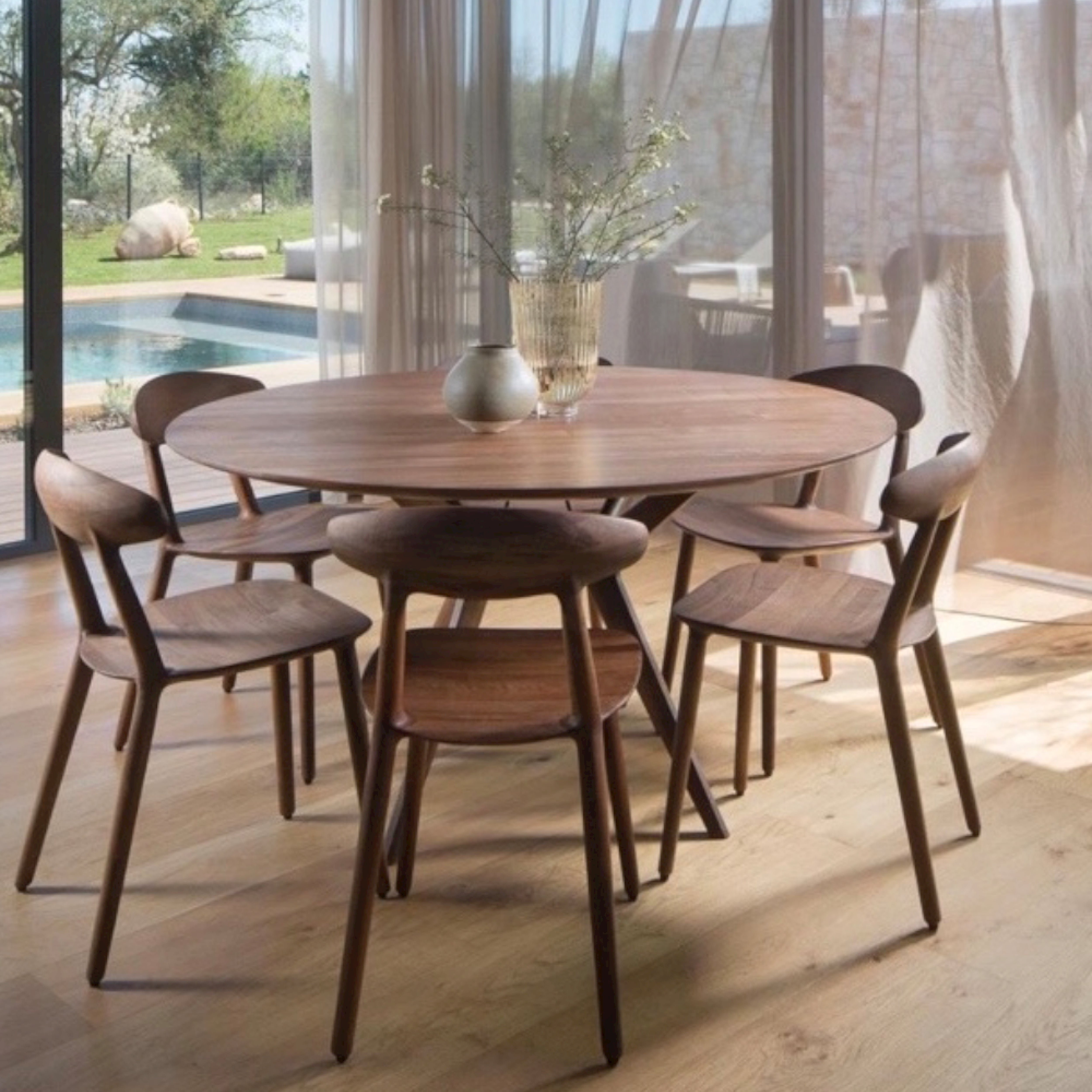 A round dining table of solid wood, with five Artisan WU Chairs, adorns a bright room. Two vases with plants rest on the table. Large windows showcase an outdoor pool and trees, as sheer curtains filter in natural light to highlight the eco-friendly materials used throughout.