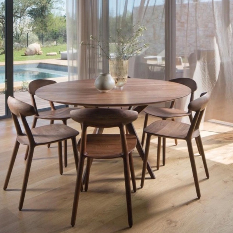 A round dining table of solid wood, with five Artisan WU Chairs, adorns a bright room. Two vases with plants rest on the table. Large windows showcase an outdoor pool and trees, as sheer curtains filter in natural light to highlight the eco-friendly materials used throughout.