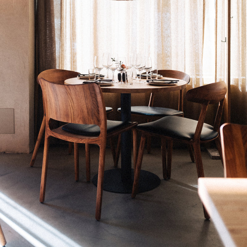A cozy dining area showcases a solid wood round table set for four with wine glasses and plates. The WU Chairs by Artisan feature black cushions, and soft natural light filters through sheer curtains, highlighting the use of eco-friendly materials.
