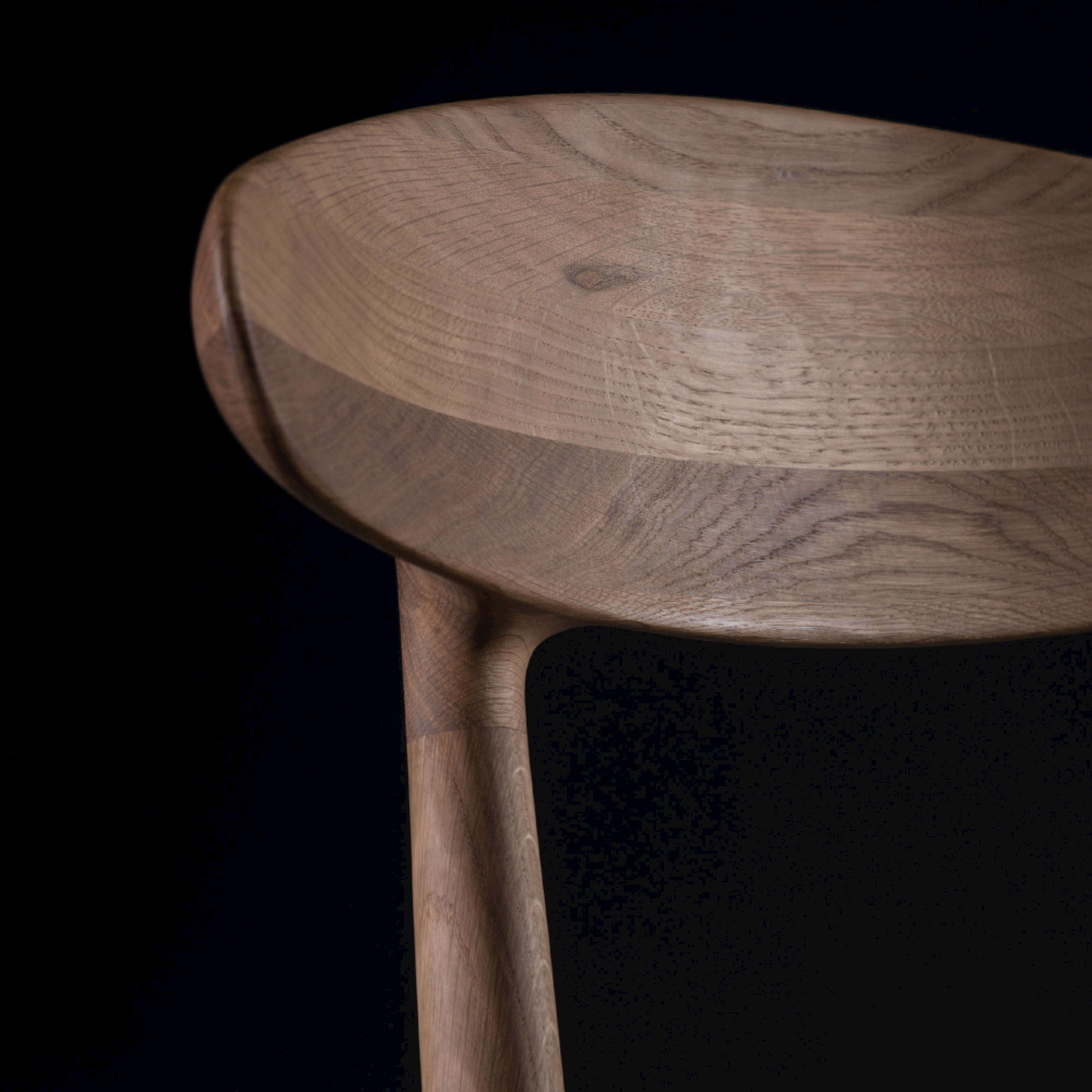The WU Chair by Artisan is featured in a close-up, highlighting the curved design and grain of its solid wood seat and backrest. Enhanced by natural oils, its smooth finish and texture stand out against a dark background.