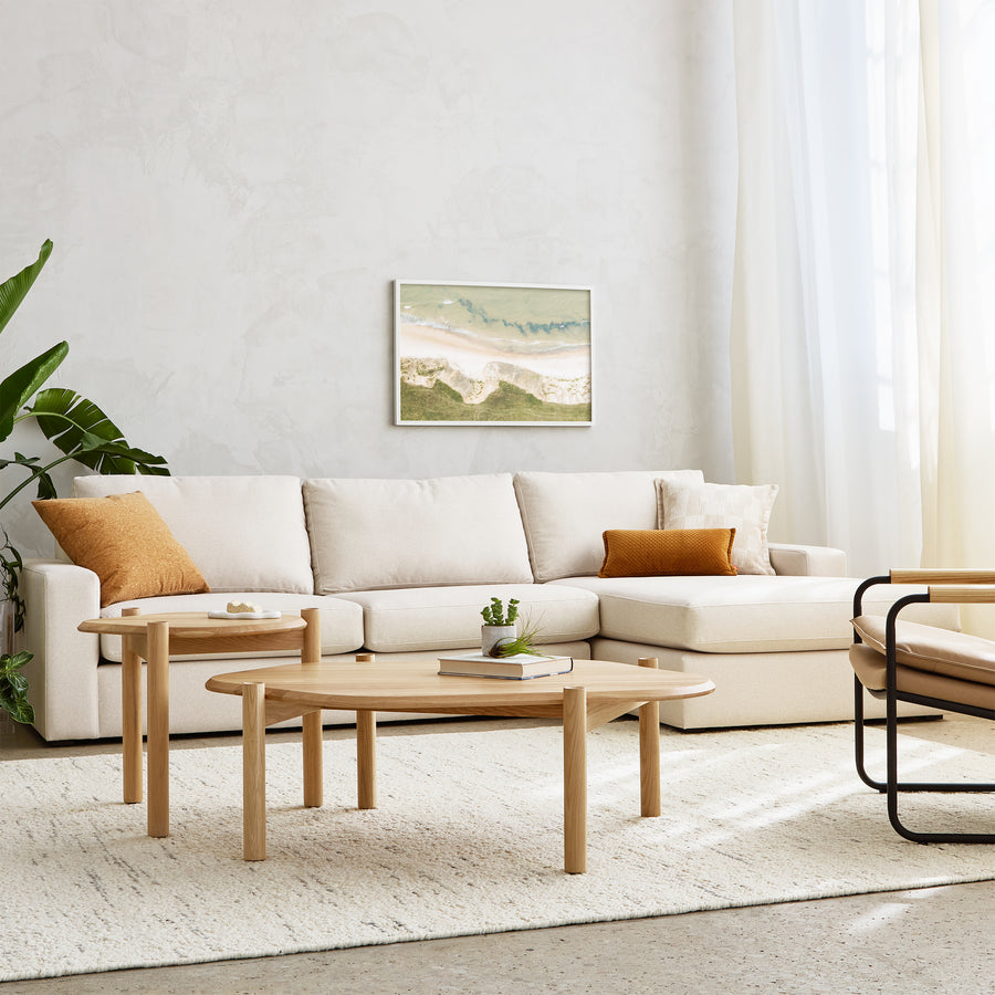 A modern living room with the Gus Modern Ashdale Right-Facing Sectional, hardwood frame, synthetic-down cushions, two wood coffee tables, tan accent pillows, potted plant, neutral rug, beach landscape art on pale walls and streaming sunlight.