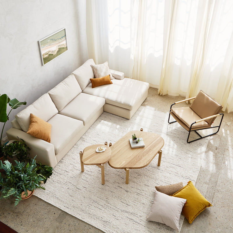 A bright, modern living room with the Gus Modern Ashdale Right-Facing Sectional featuring synthetic-down cushions, a tan accent chair, nested wood coffee tables, potted plants, decorative pillows, and a textured rug by sheer-curtained windows.