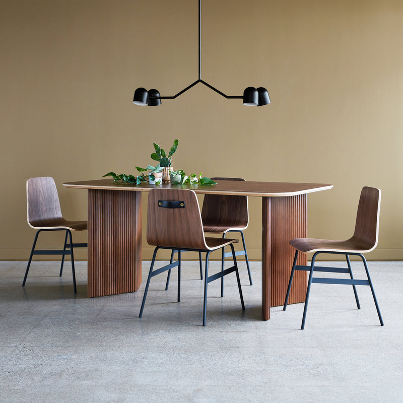 A modern dining area with the Atwell Dining Table Rectangle by Gus Modern, four wooden chairs with black metal legs, a minimalist black chandelier, and green plants on the tabletop, set against a beige wall and concrete floor.