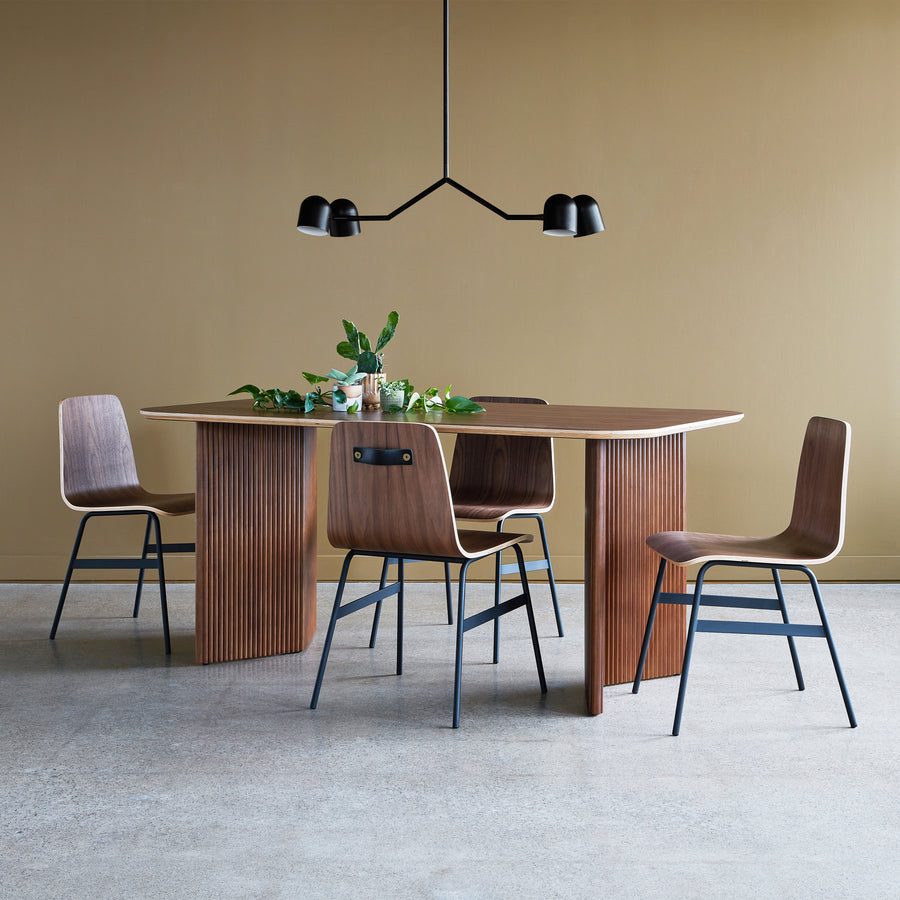 A modern dining area with the Atwell Dining Table Rectangle by Gus Modern, four wooden chairs with black metal legs, a minimalist black chandelier, and green plants on the tabletop, set against a beige wall and concrete floor.