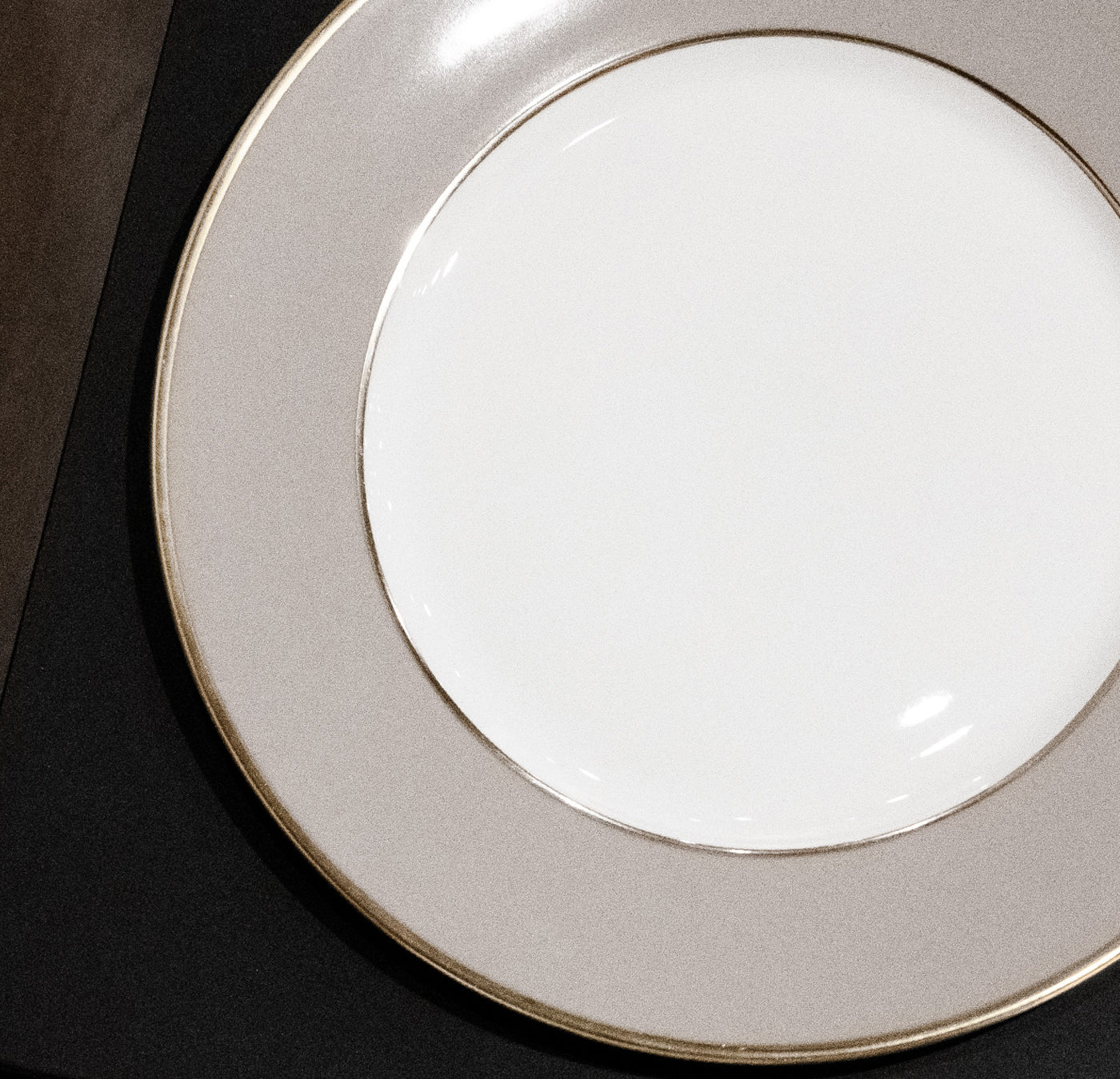 A close-up of the Serving Plate Gold Rim by Augarten on a dark surface showcases its white porcelain with a wide, light gray rim, elegantly hand-painted for an artistic touch.