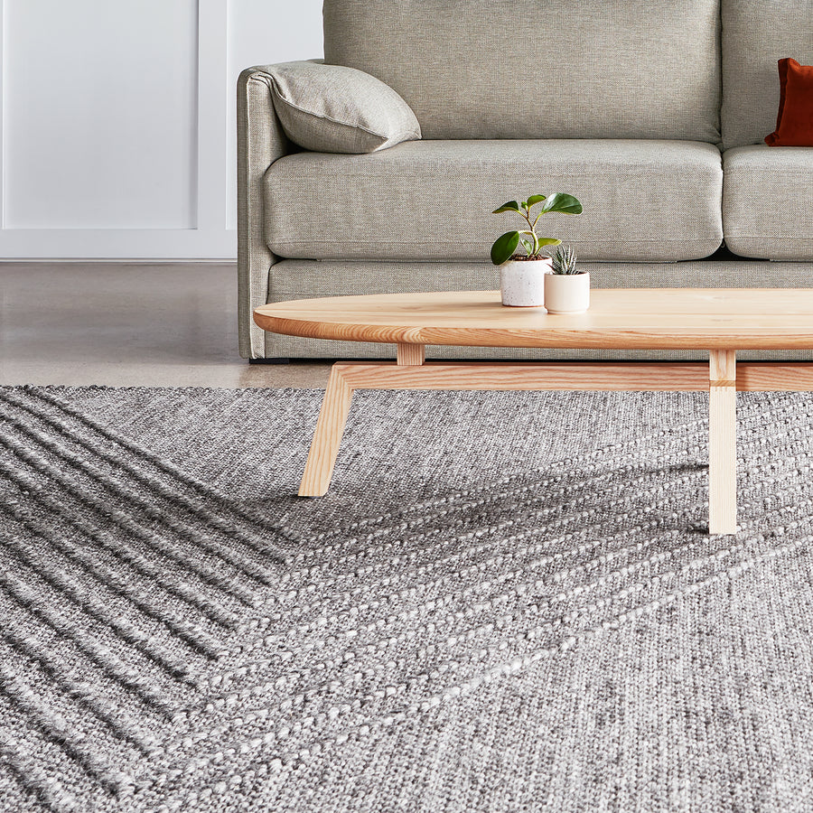 A light wooden coffee table with two small potted plants sits on the Avro Rug by Gus Modern in front of a beige sofa with a cushion in a modern living room.