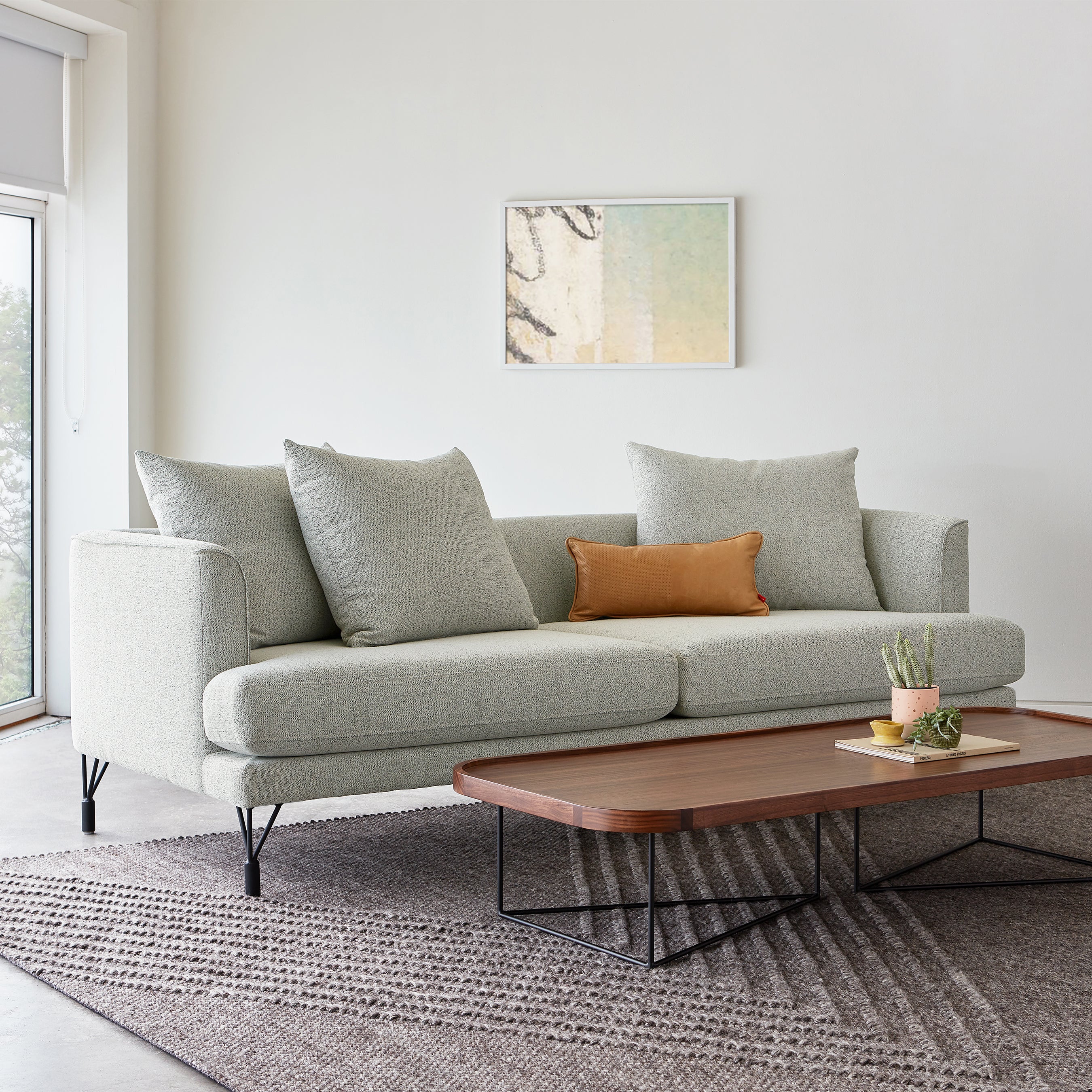 A modern living room with a light gray sofa, a rectangular wooden coffee table with metal legs, a small plant, and abstract art on the wall features the Avro Rug by Gus Modern, hand-knotted from New Zealand wool for added texture and warmth.