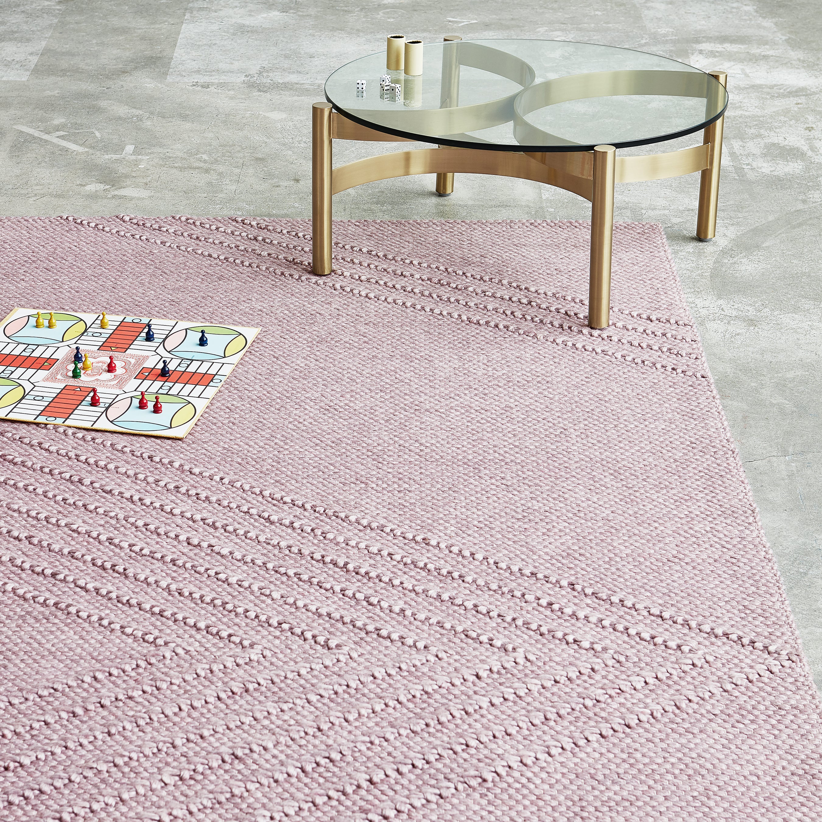 The Avro Rug by Gus Modern, a hand-knotted textured pink rug featuring a Ludo board game and pieces, sits beside a round glass coffee table with gold legs on a light gray tiled floor.