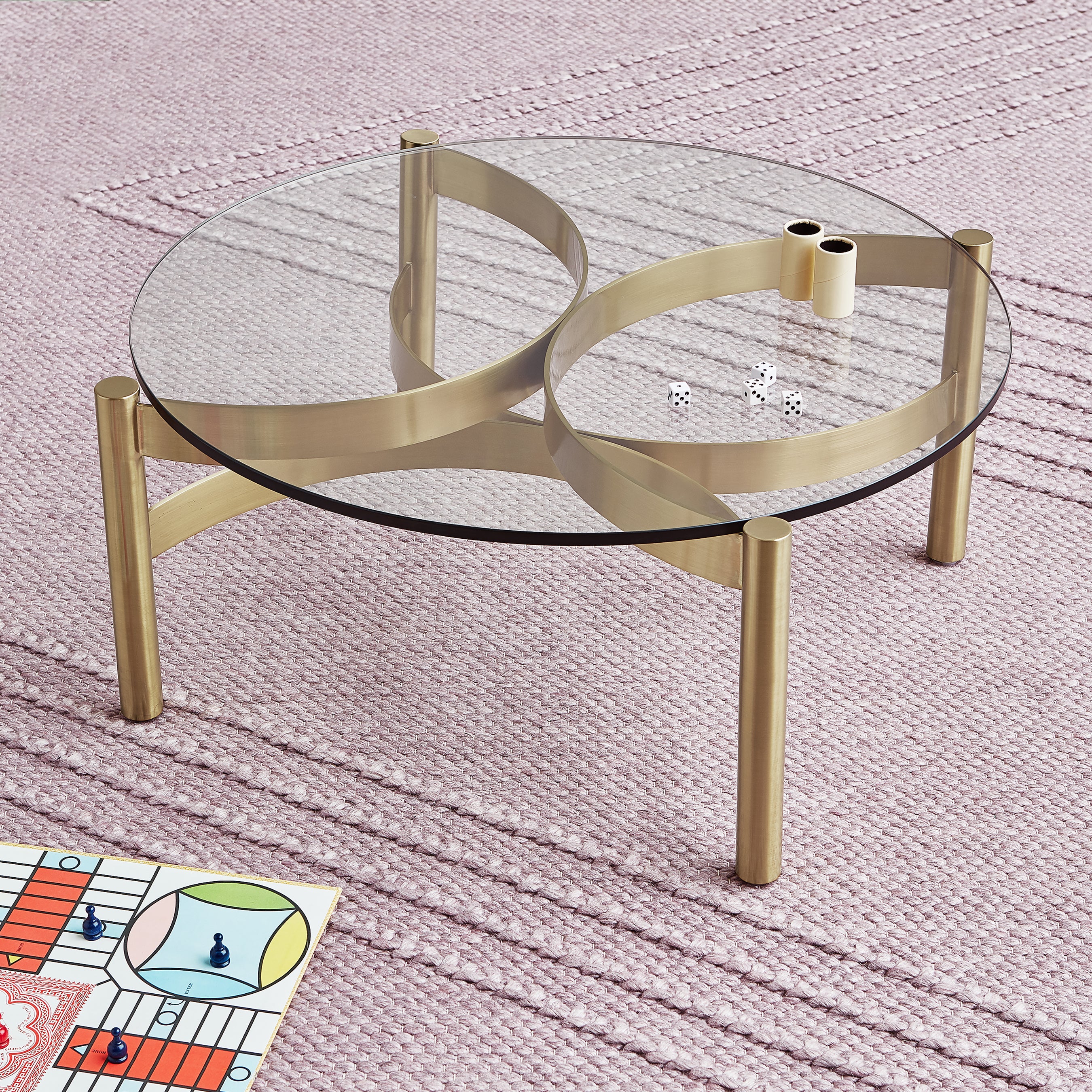 A round glass coffee table with a gold frame sits on a textured pink Avro Rug by Gus Modern. Two dice and game pieces rest on the table, with a colorful board game partially visible in the lower left corner.