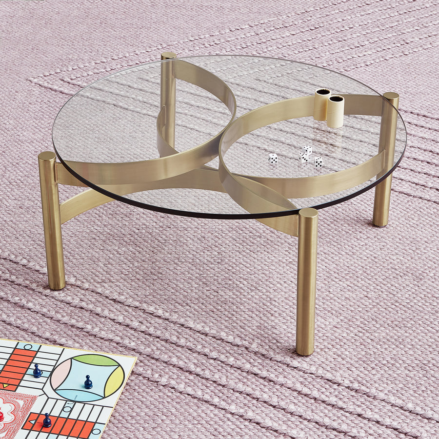 A round glass coffee table with a gold frame sits on a textured pink Avro Rug by Gus Modern. Two dice and game pieces rest on the table, with a colorful board game partially visible in the lower left corner.