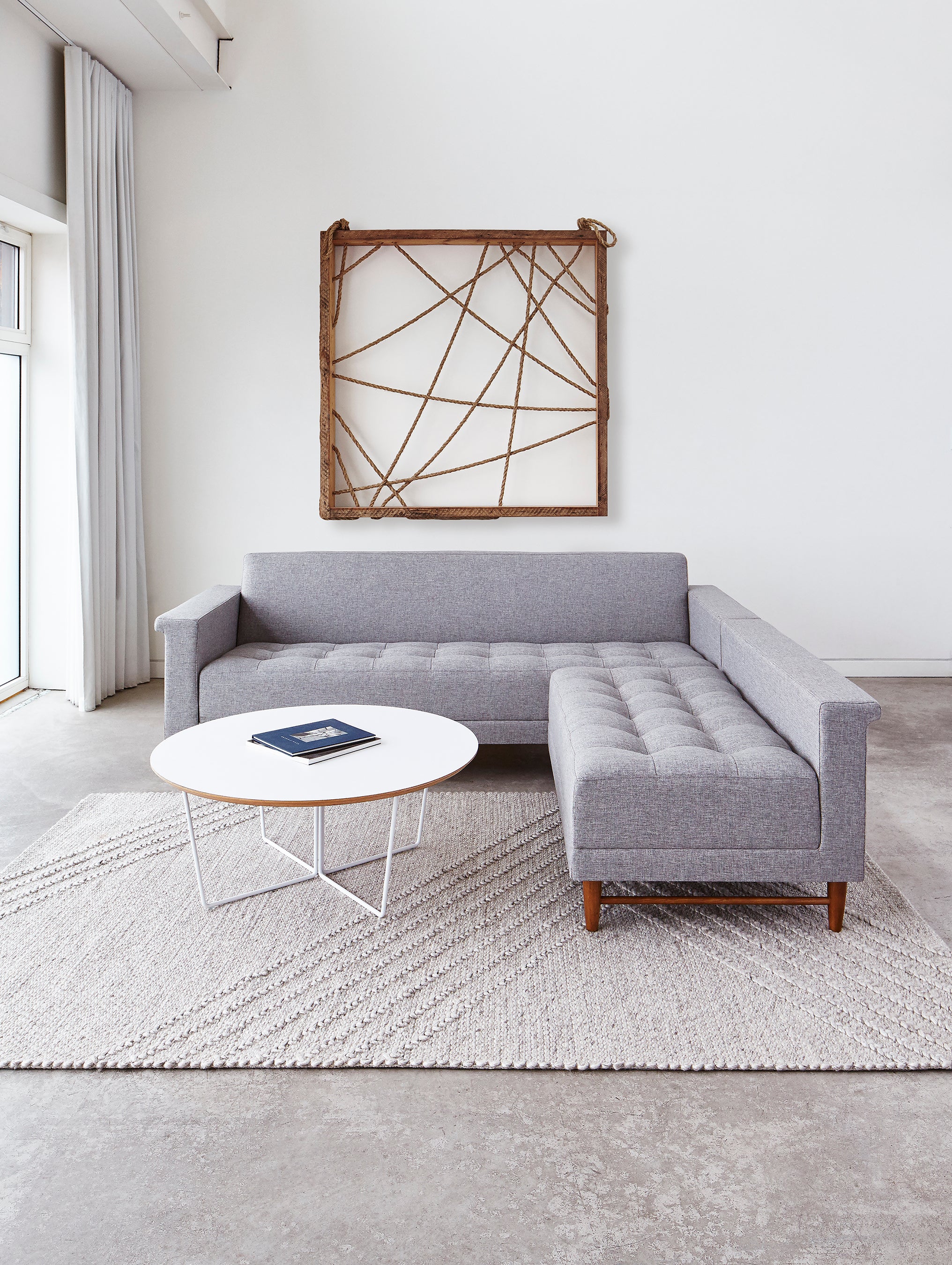 A modern living room with a gray sectional sofa, a round white coffee table topped with a book, and abstract wooden wall art on white walls. The Gus Modern Avro Rug adds texture, while natural light streams in from large windows on the left.