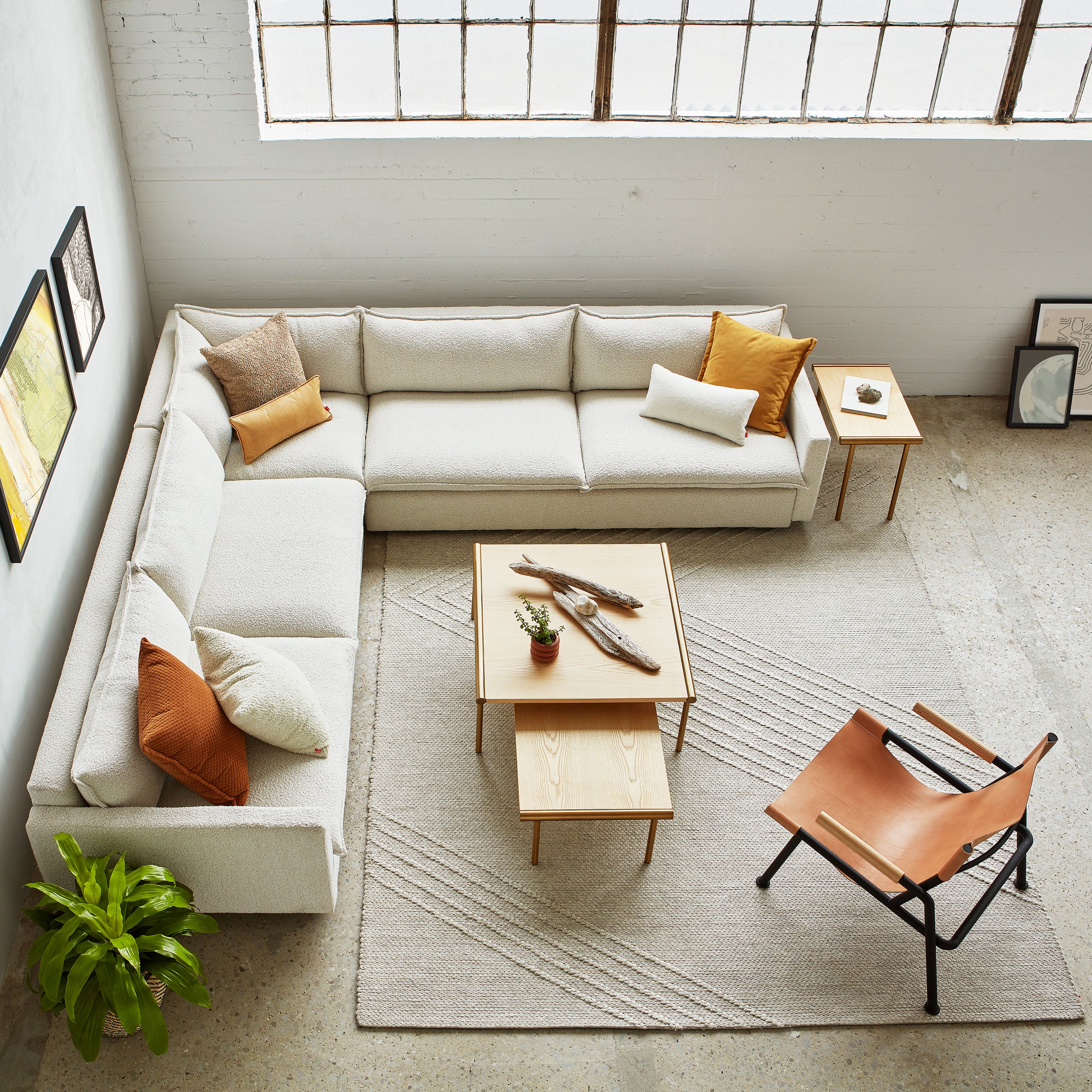 A modern living room with an L-shaped beige sofa, orange and white pillows, a tan accent chair, wooden tables, potted plants, and framed wall art on light walls and floors is finished with the minimalist Avro Rug by Gus Modern for a stylish touch.