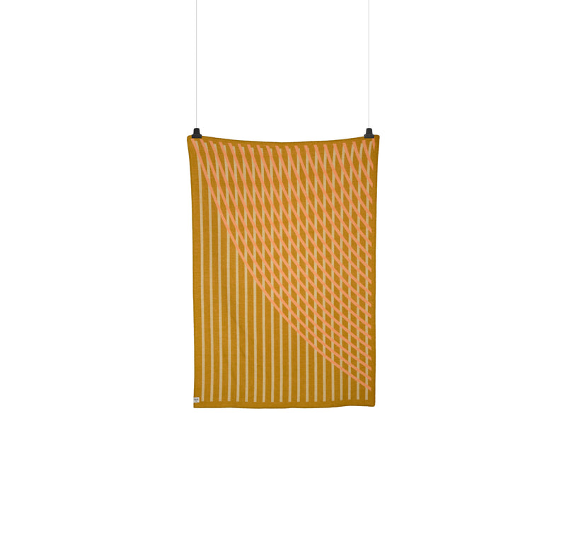 The BISLETT Throw by Roros is a vertically hanging textile made of Norwegian lambswool, with a geometric pattern featuring diagonal lines in mustard yellow and soft pink that evoke a Moiré wave effect against a plain white background.
