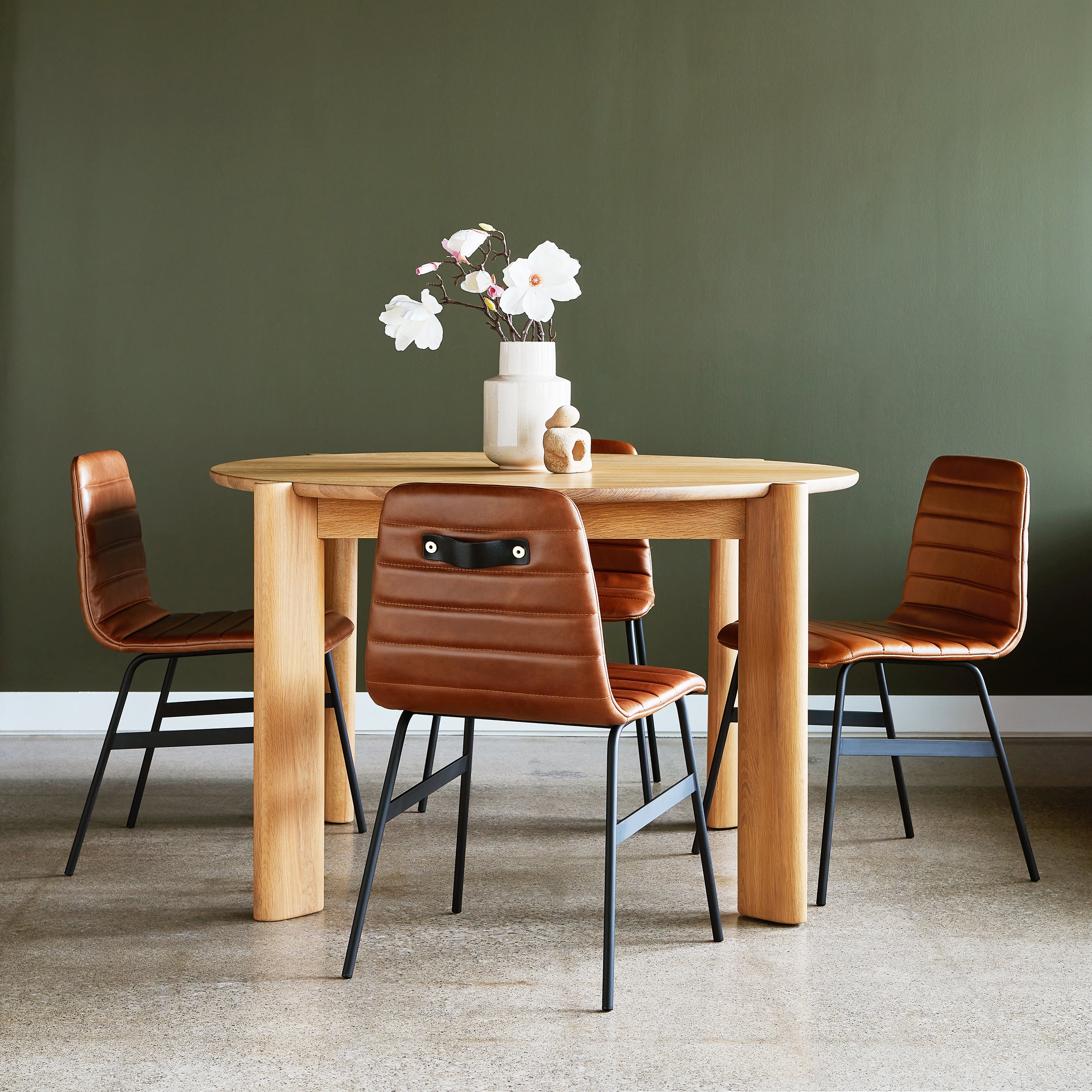 A Gus Modern Bancroft Dining Table Round, paired with four brown leather chairs, sits by a green wall on polished concrete. The modern table is topped with a white vase of flowers and two ceramic pieces.