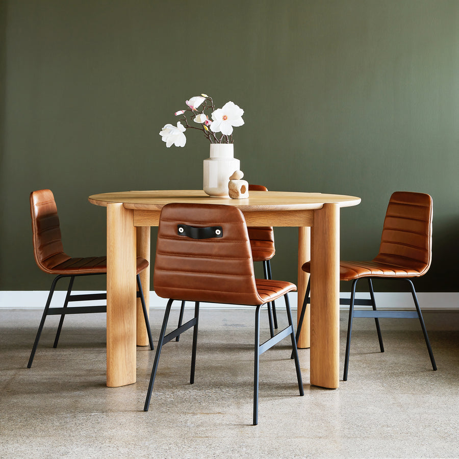 A Gus Modern Bancroft Dining Table Round, paired with four brown leather chairs, sits by a green wall on polished concrete. The modern table is topped with a white vase of flowers and two ceramic pieces.