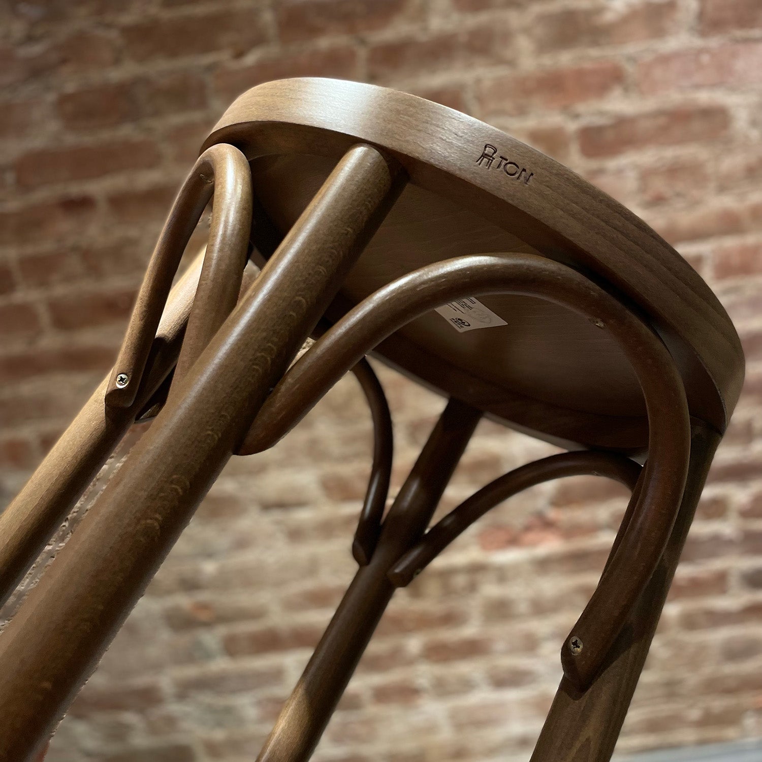 The Barstool 73 by TON, visible from a low angle against a brick wall, boasts beech leg joints and curved legs. It features an upholstered seat with a smooth, polished finish and sturdy appearance.