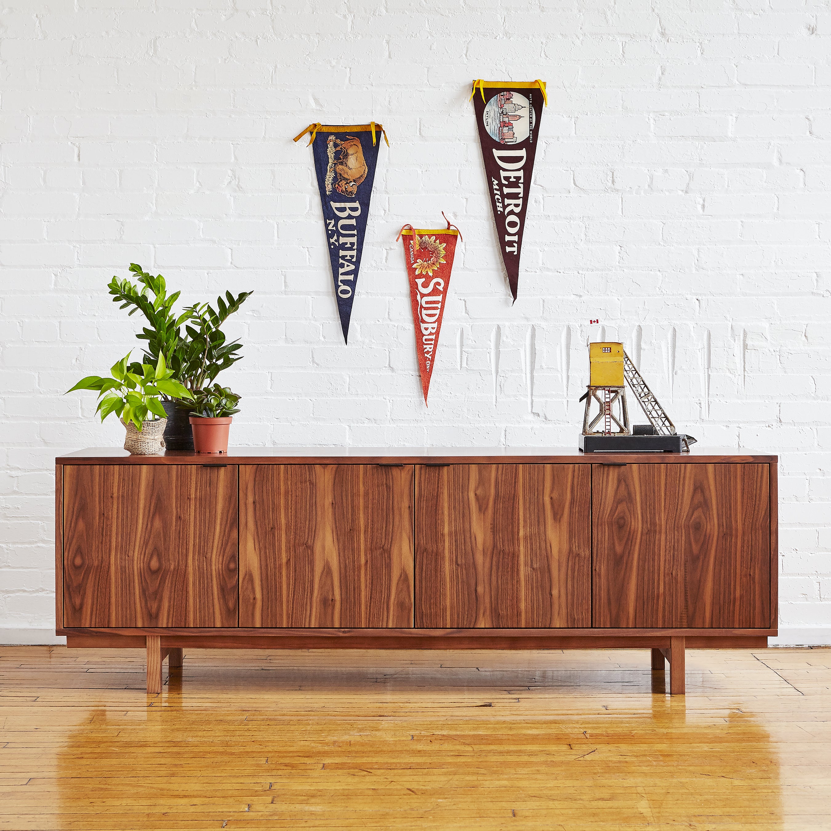 The Gus Modern Belmont Credenza stands against a white brick wall, topped with potted plants. Above it hang vintage pennants for Buffalo, Detroit, and Sudbury, along with a small model oil rig structure.