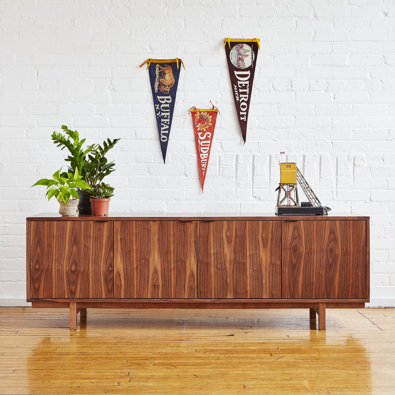 The Gus Modern Belmont Credenza stands against a white brick wall, topped with potted plants. Above it hang vintage pennants for Buffalo, Detroit, and Sudbury, along with a small model oil rig structure.