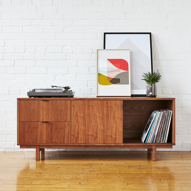 The Gus Modern Belmont Media Stand sits against a white brick wall, displaying a turntable, framed abstract art, a small plant, and vinyl records. A shiny wooden floor enhances this stylish mid-century modern storage cabinet.
