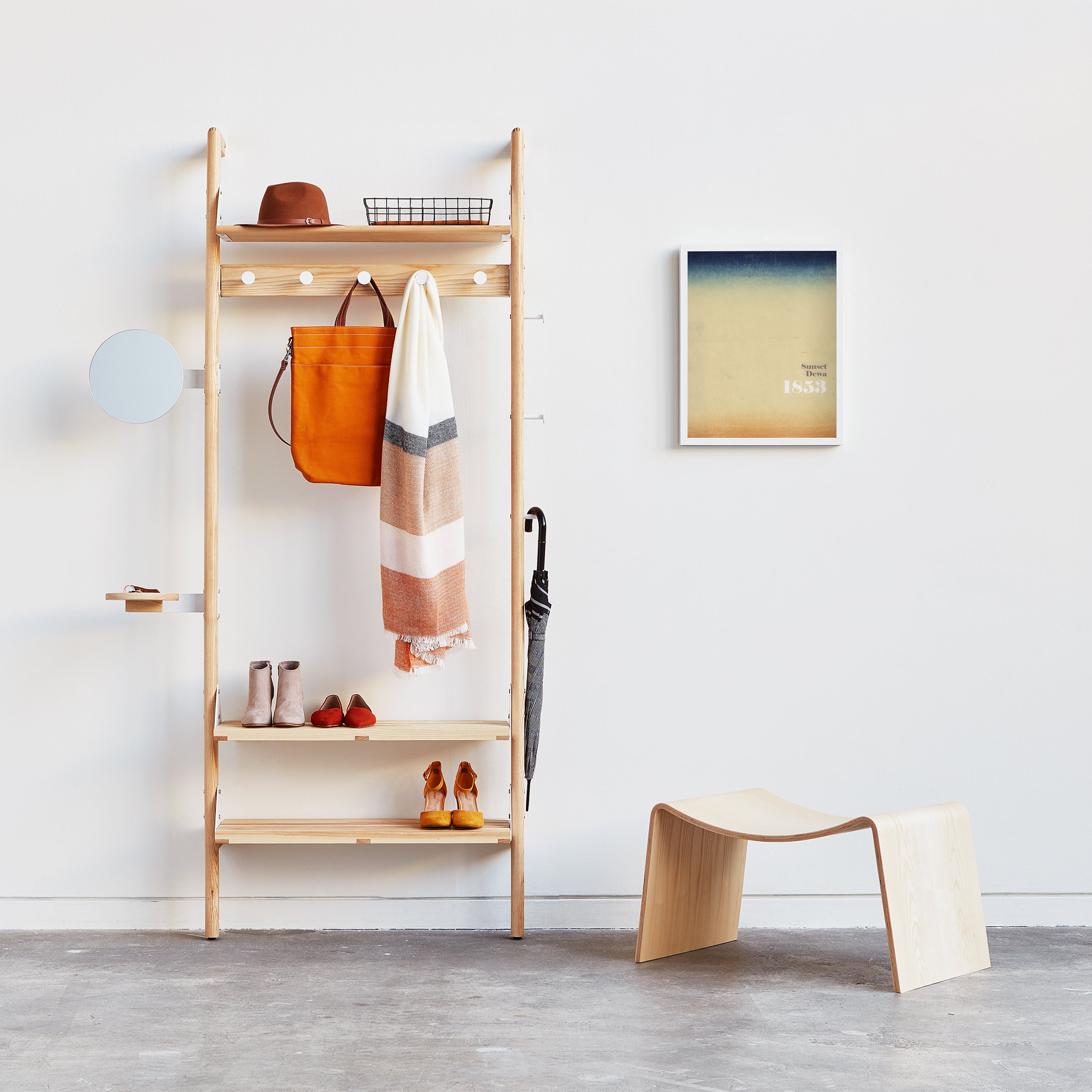 The Branch 1 Entryway Unit by Gus Modern features customizable shelves for hats, scarves, bags, shoes, and umbrellas. A round mirror is mounted on the left, with abstract wall art and a light wooden bench nearby against a white wall.