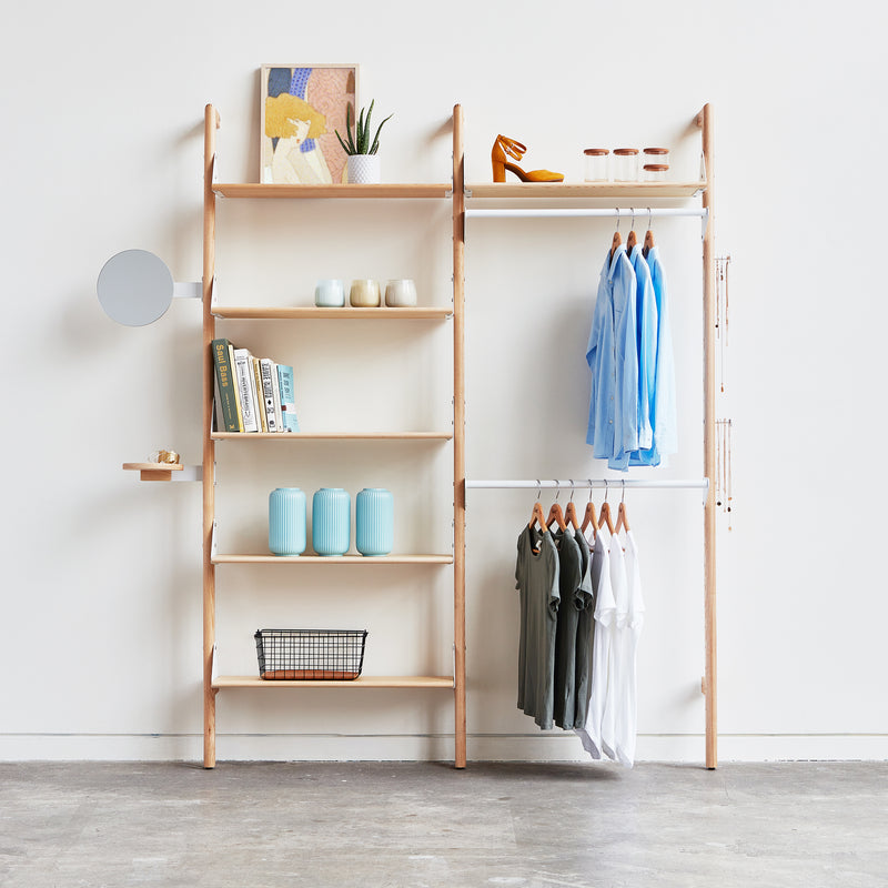 The Branch 2 Display Unit by Gus Modern, a minimalist wooden shelving piece, offers open storage for books, jars, plants, décor, and shirts against a white wall—complete with shoes and a round mirror for versatile display.
