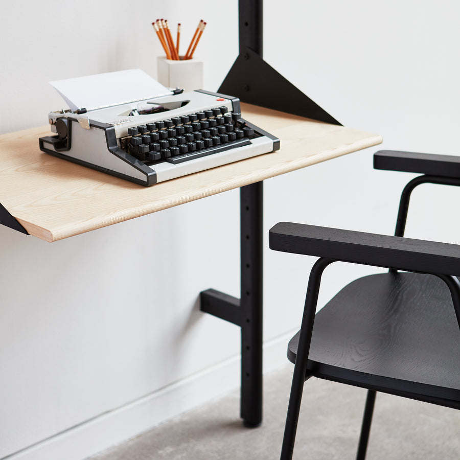 A modern workspace features a black chair, light wood floating desk, and vintage typewriter, accented by Gus Modern’s Branch Desk Shelving Unit Add-On in mid-century style against a white wall, with pencils stored neatly nearby.