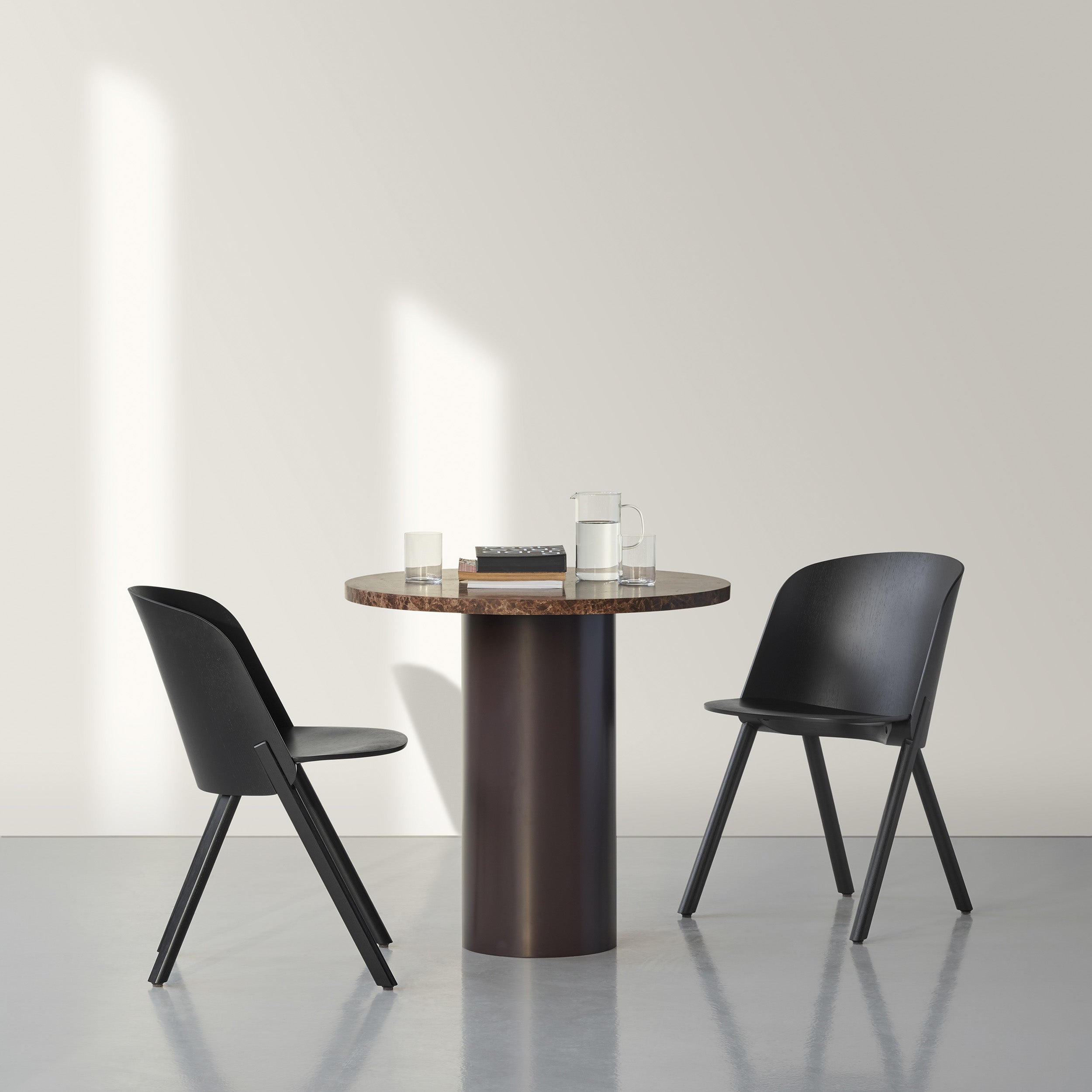 A minimalist room showcases an E15 CT09 ENOKI BISTRO table with a round wooden surface and two black chairs. The table displays a jug, two glasses, and stacked books. Soft sunlight casts shadows on the neutral-colored wall and floor.