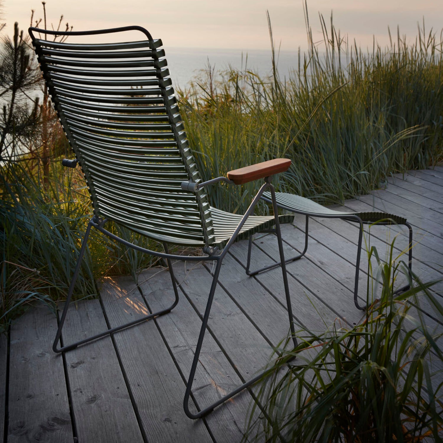 The Houe Click Position Chair, a weather-resistant metal lounge chair with wooden armrests, rests on a wooden deck amid tall grass, offering views over a serene body of water under a hazy sunset sky.