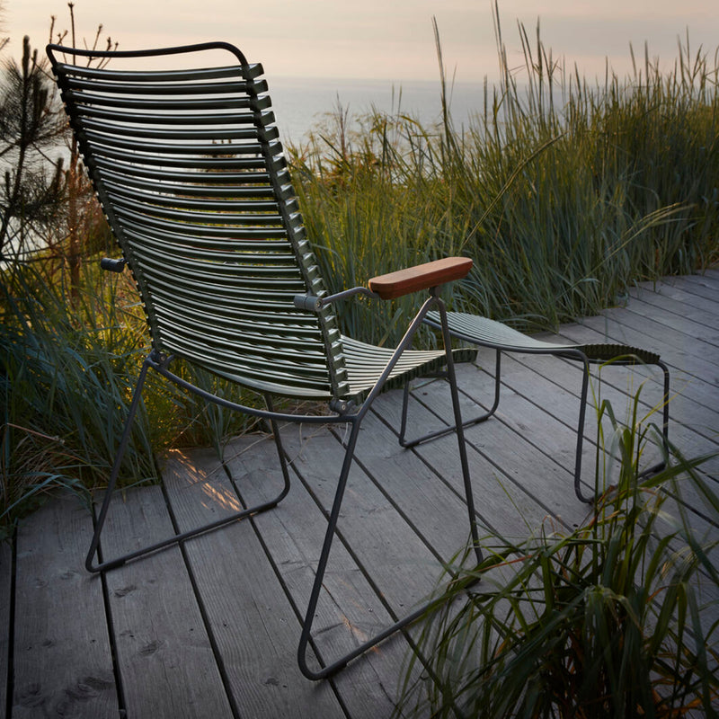 The Houe Click Position Chair, a weather-resistant metal lounge chair with wooden armrests, rests on a wooden deck amid tall grass, offering views over a serene body of water under a hazy sunset sky.