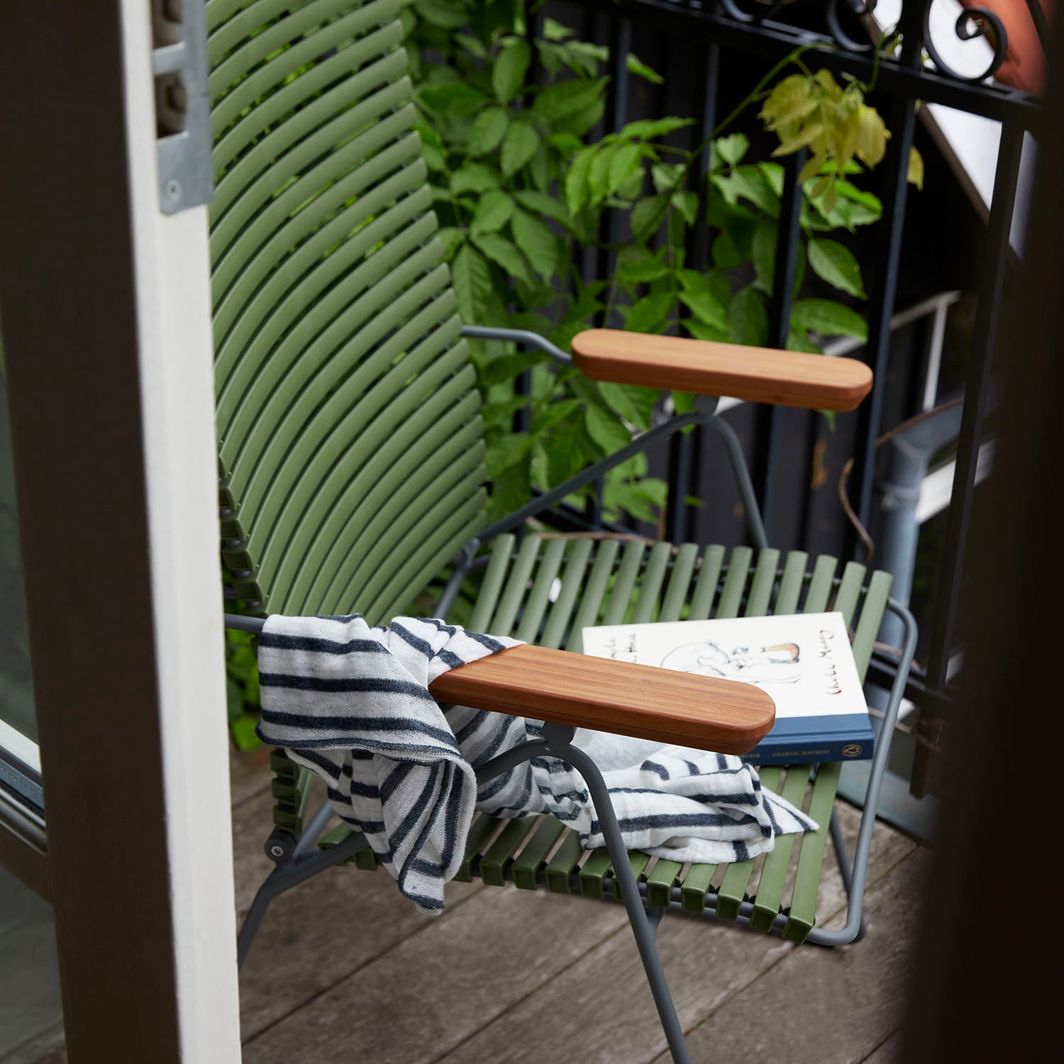 The Houe Click Position Chair, designed for outdoor use, sits on the balcony. Adorned with a striped black and white towel and a book, it is surrounded by leafy plants near the black railing, visible through an open door.