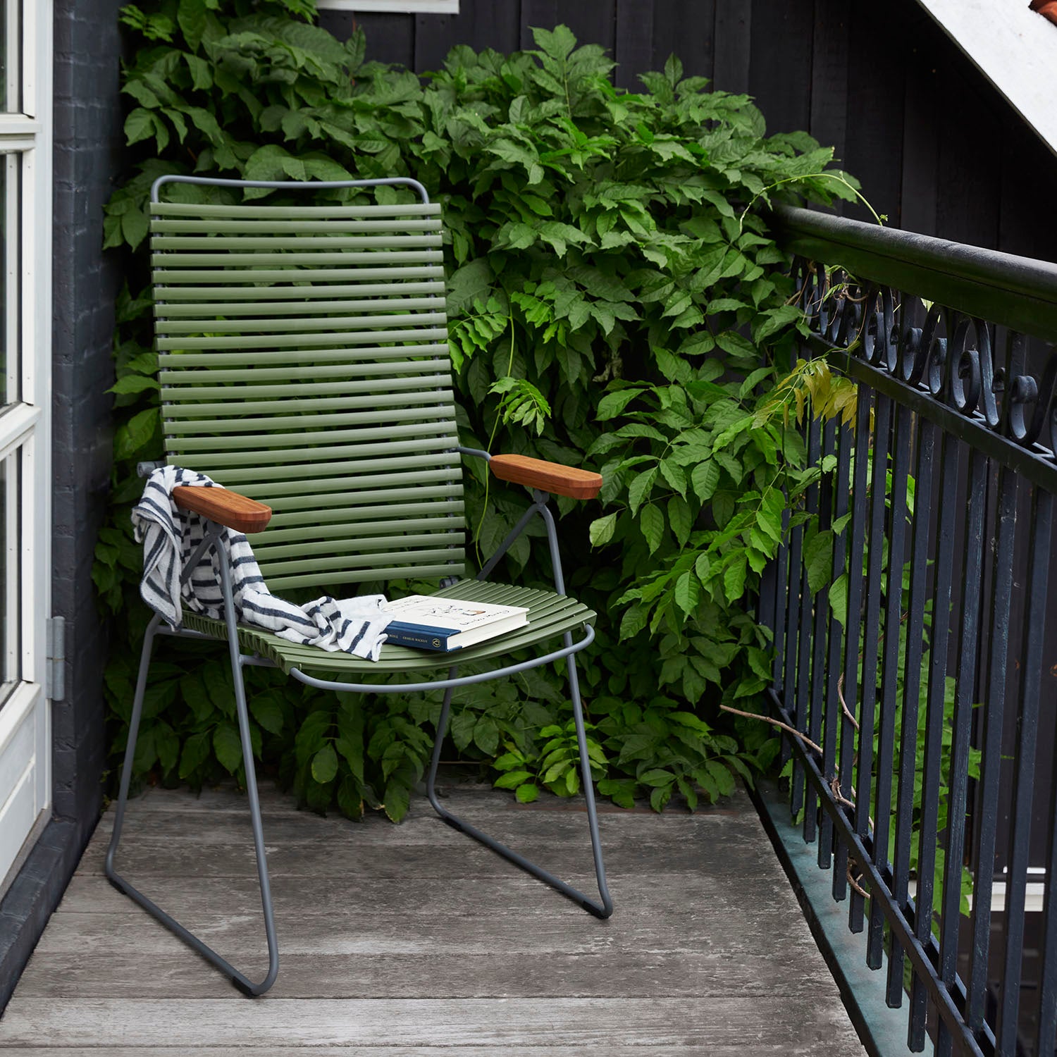 A Click Position Chair by Houe, featuring green stripes and weather-resistant material with wooden armrests, is on a deck surrounded by lush plants. A striped fabric and book are placed on the chair. The balcony railing to the right adds to this cozy retreat.
