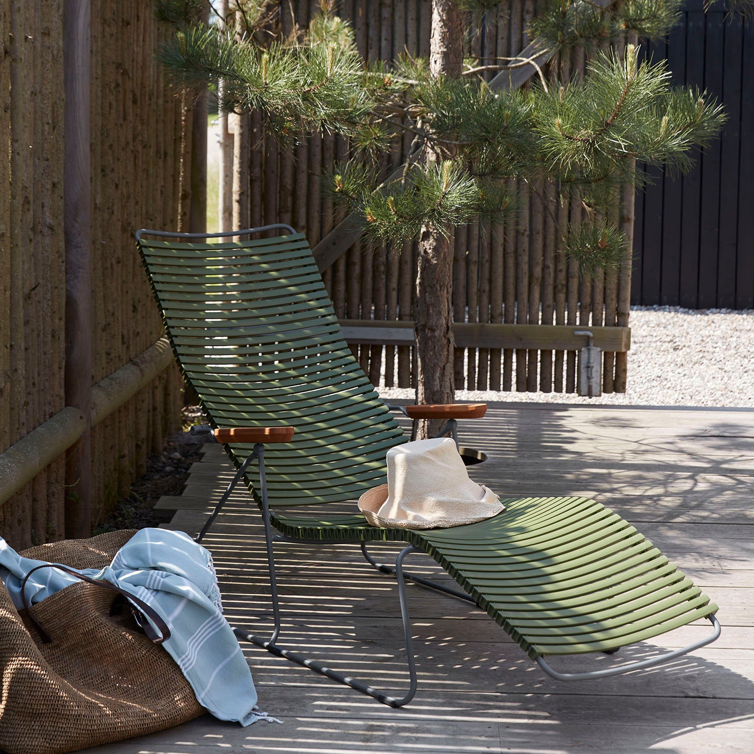 The Houe Click Sunlounger, featuring a green ribbed design and wooden armrests, sits on a stone patio under a small tree for UV-protected relaxation. A hat rests on the lounger. Nearby, a blue blanket and brown bag are nestled beside a bamboo fence in this welcoming outdoor space.
