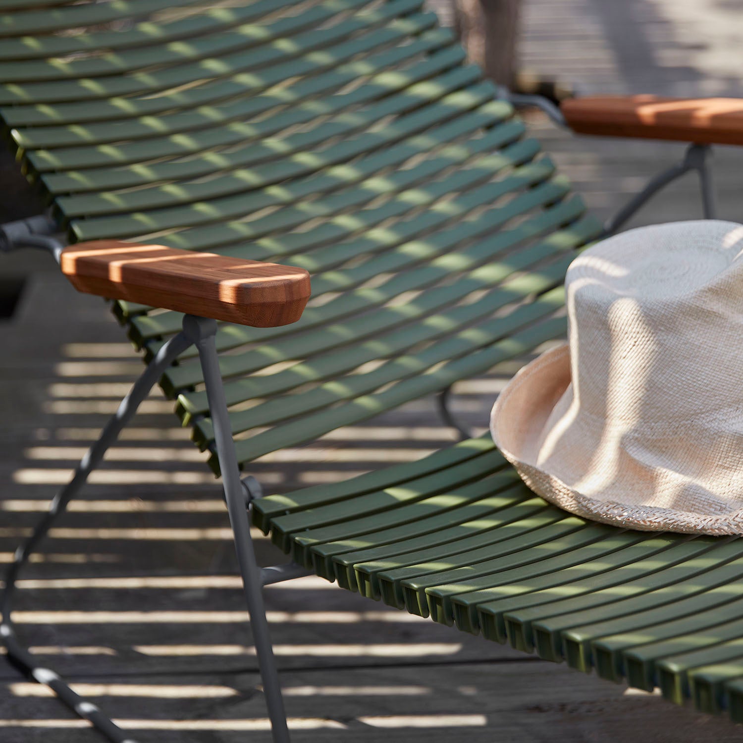Close-up of a UV-protected Click Sunlounger by Houe in green with wooden armrests. A straw hat rests on the seat, casting a shadow. Sunlight highlights the texture of this cushion-free option, surrounded by a wooden deck.